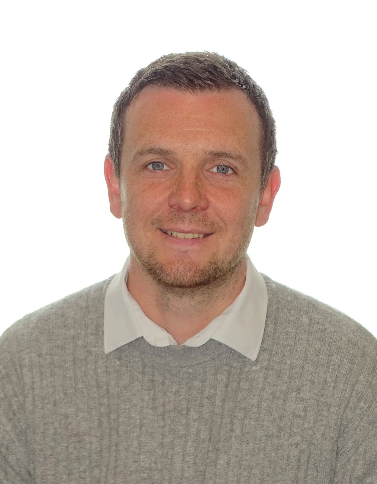 A man with short brown hair, blue eyes, and a short beard, wearing a light grey sweater over a white collared shirt, smiling, with a plain white background.