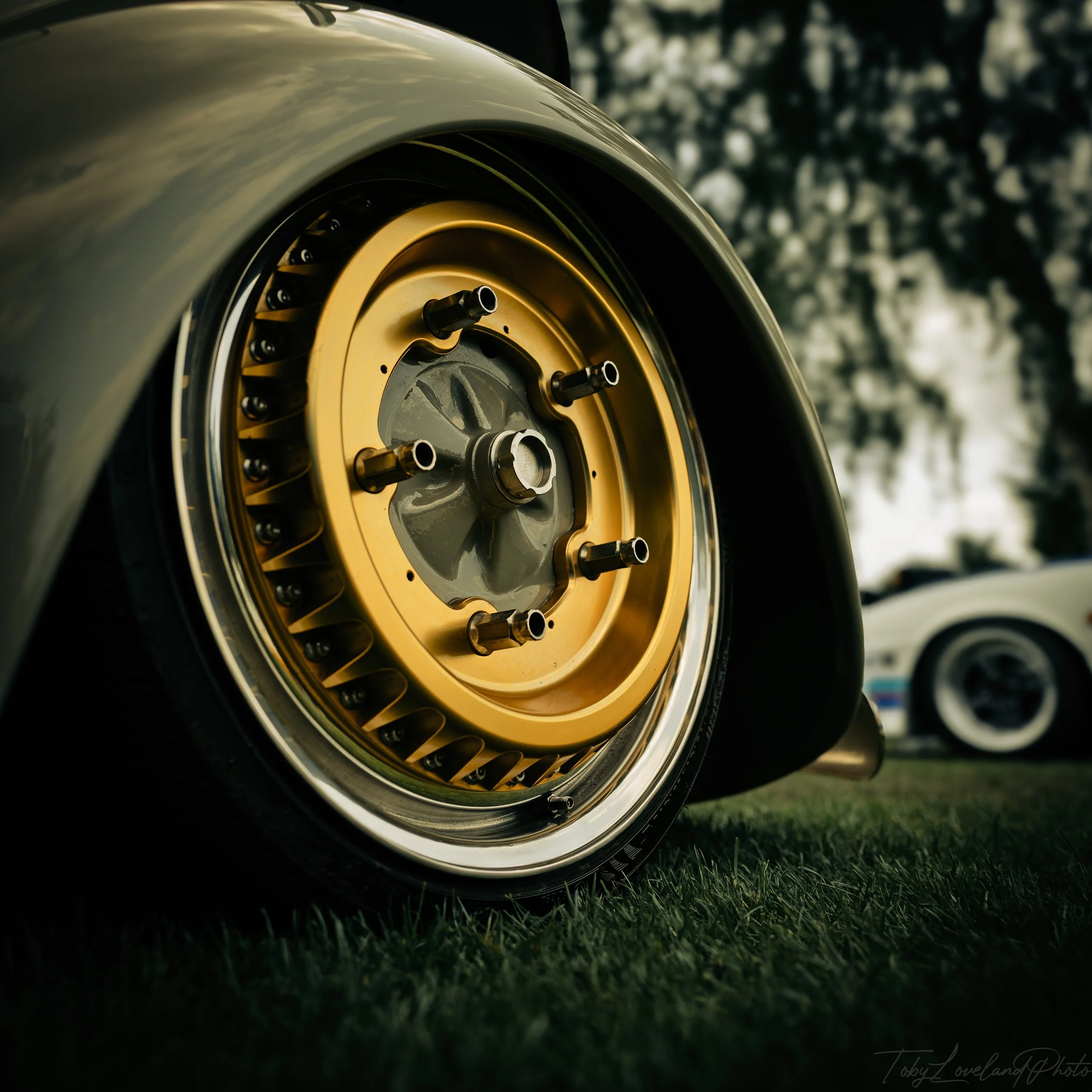 Close-up of a vintage car wheel with a gold rim and black lug nuts, on a grassy surface with blurred trees and another car in the background.