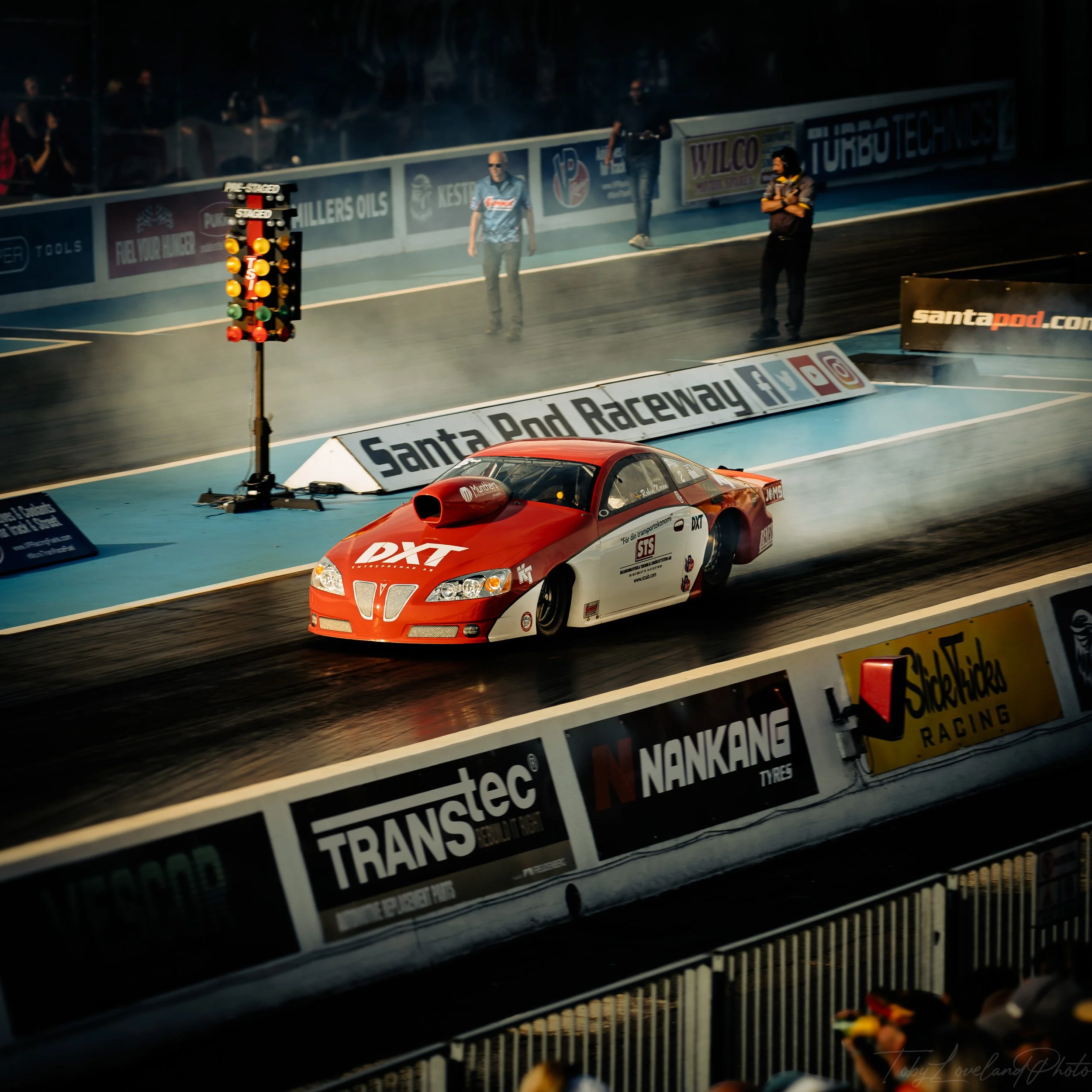 A red and white drag racing car on the track at Santa Pod Raceway, with racers and officials standing beside the track and a trackside lighting signal.