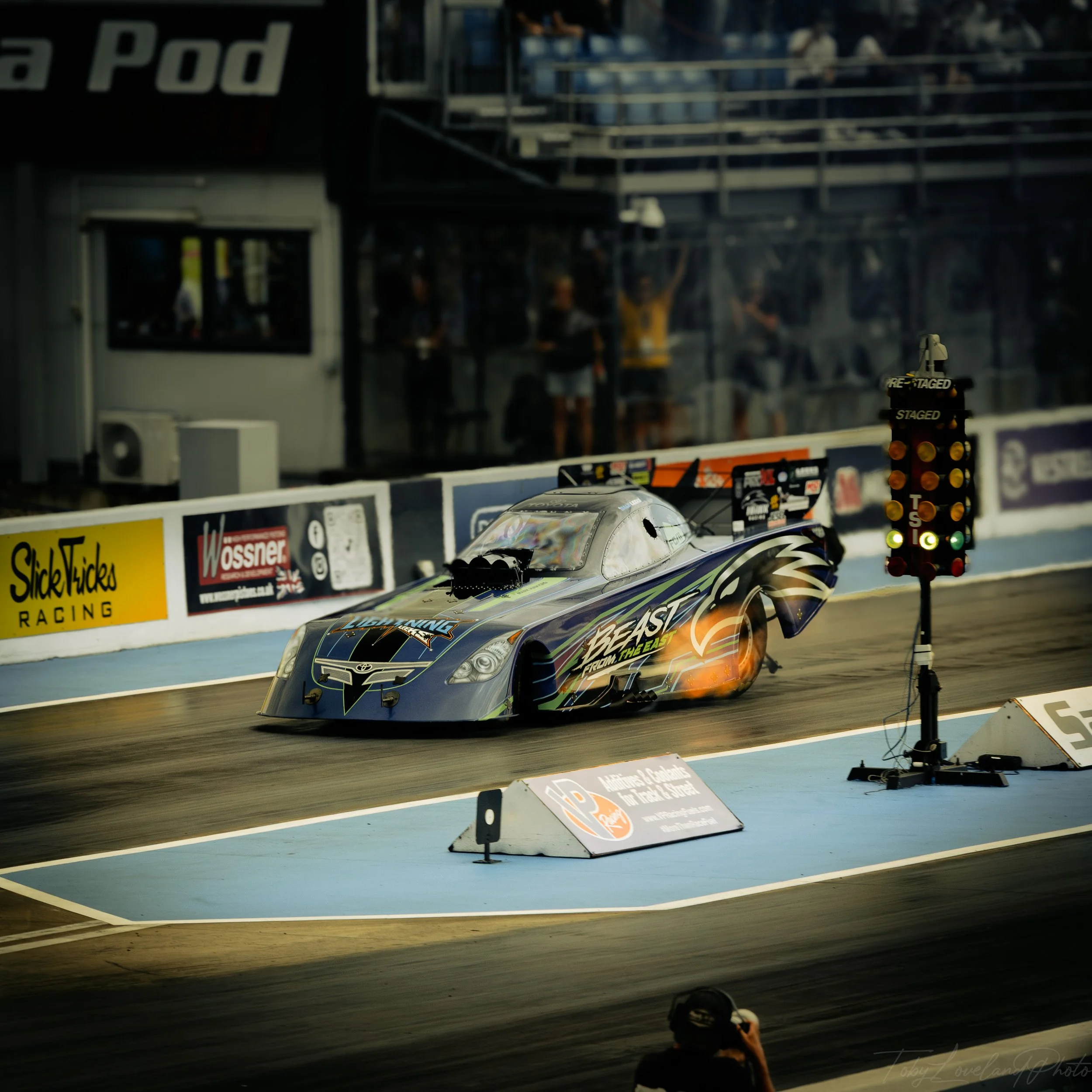 A drag racing car on the track with flames coming from the exhaust, a staging light tree beside it, and spectators in the background.