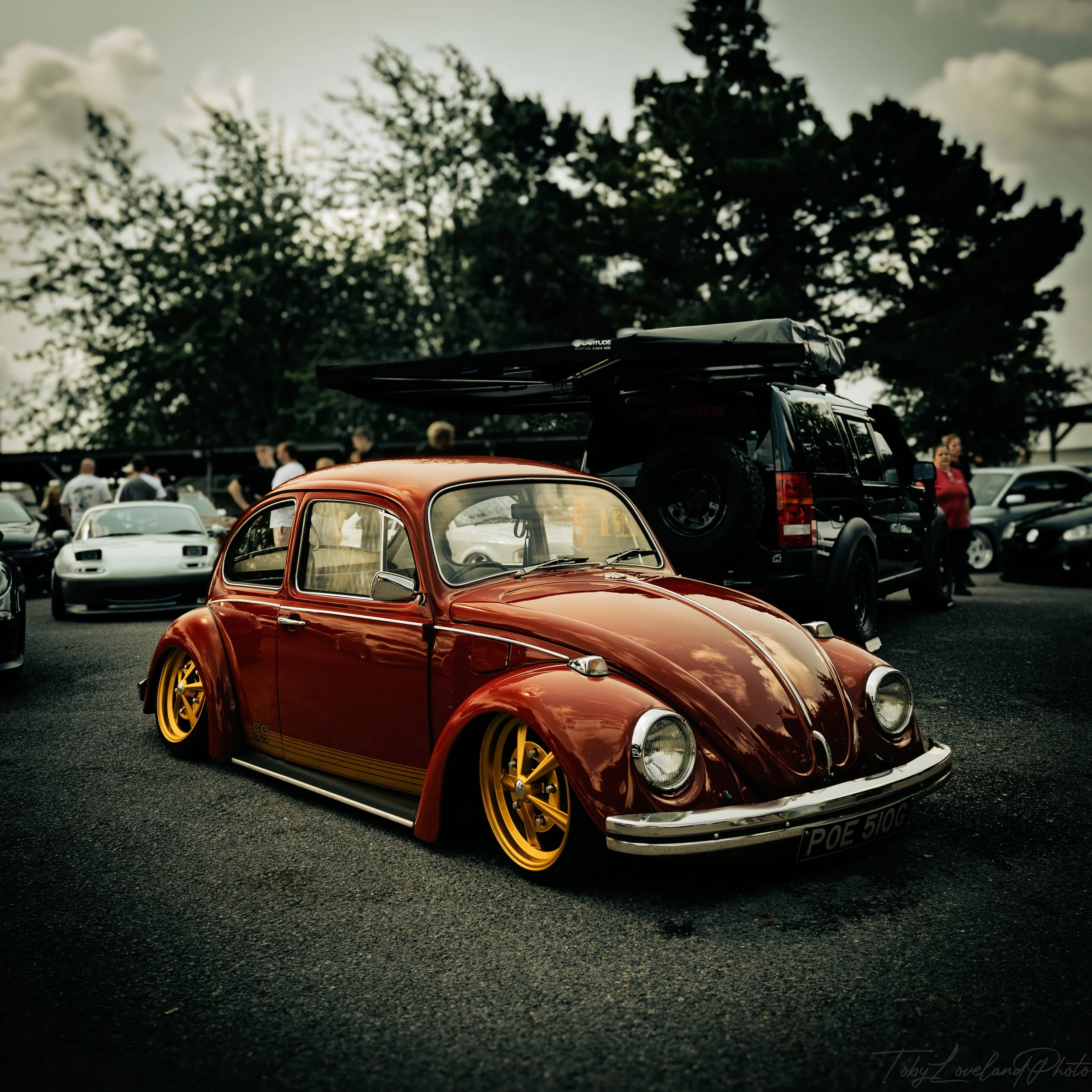 A vintage red Volkswagen Beetle with lowered stance and yellow wheels parked at a car show, surrounded by other cars and people.