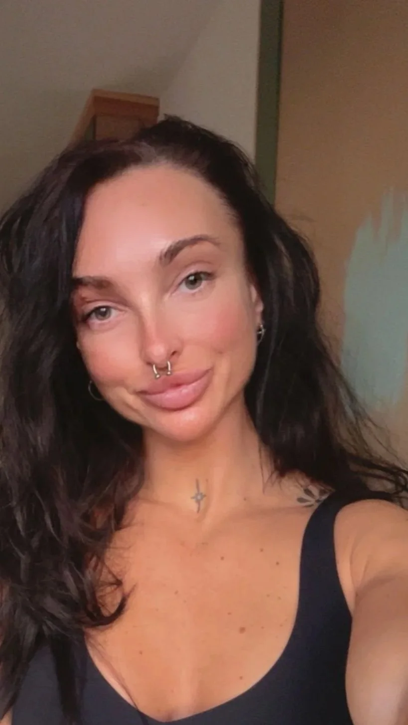 Portrait selfie of a woman with long dark hair, facial piercings, tattoos, and a black tank top indoors
