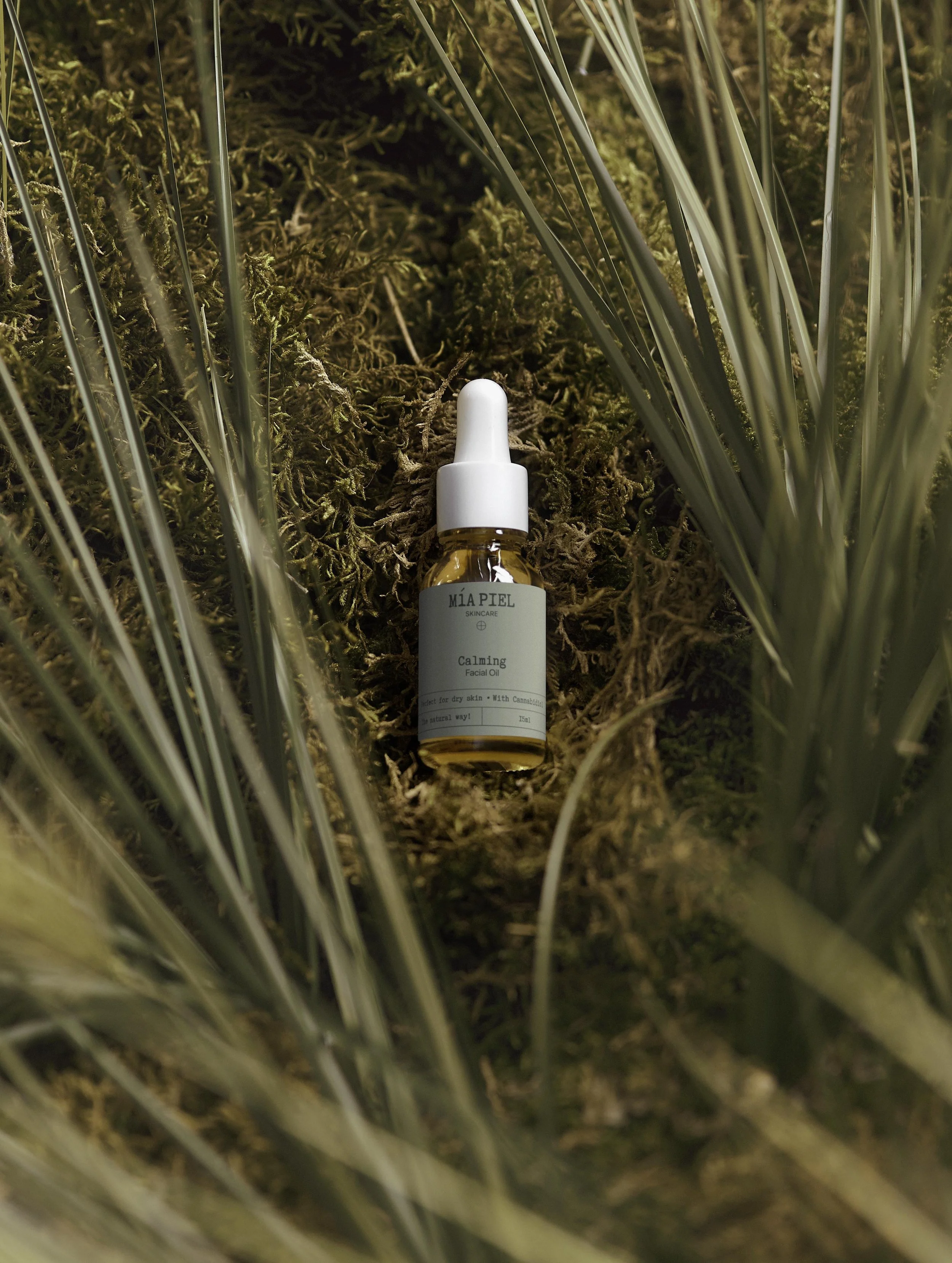 A small dropper bottle labeled 'MIA PIEL Skincare Calming Facial Oil' placed on moss among grass and foliage.
