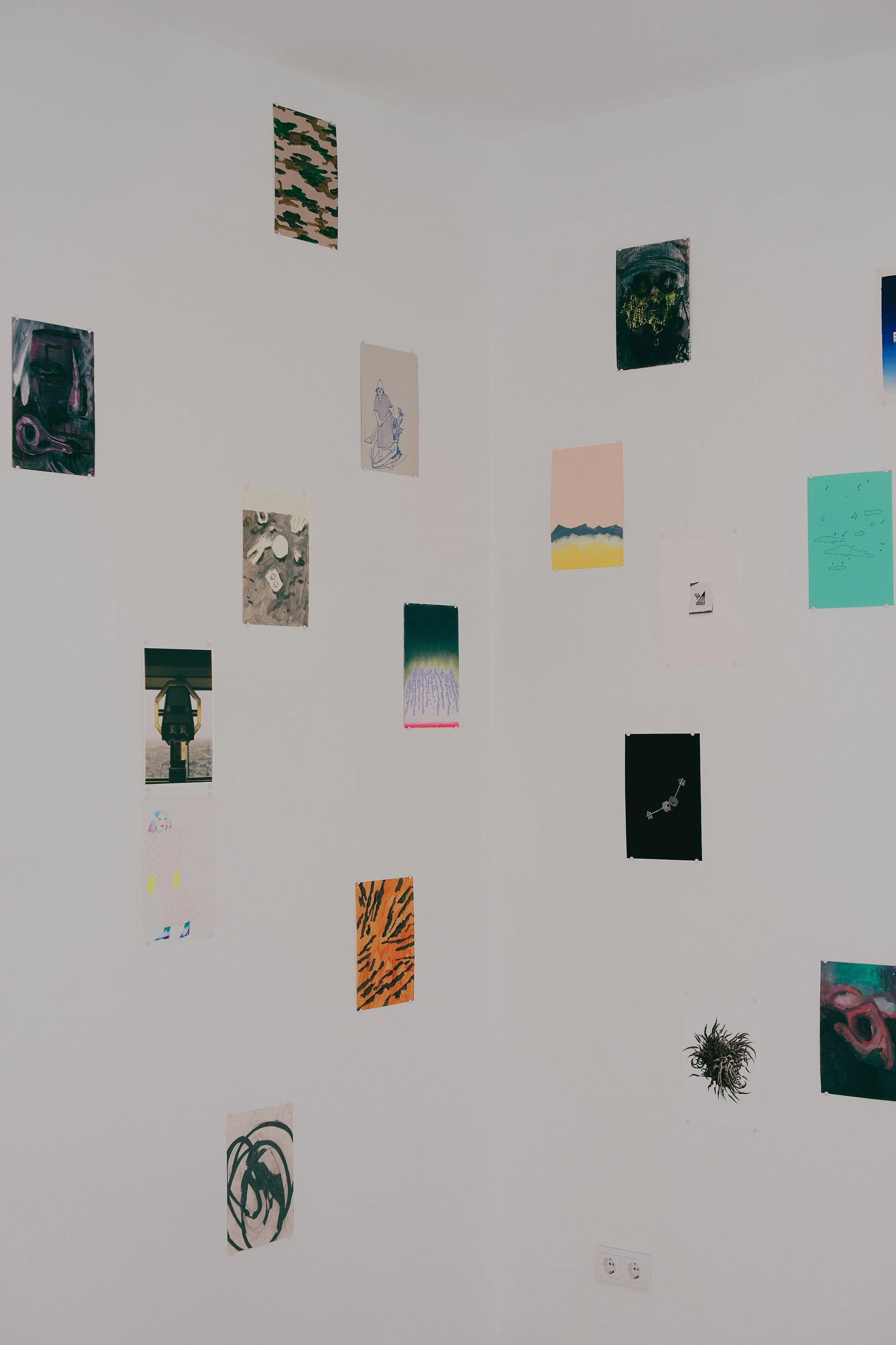 Bunte Kunstwerke hängen an einer weißen Wand, darunter eine kleine Pflanze und eine Doppelsteckdose.