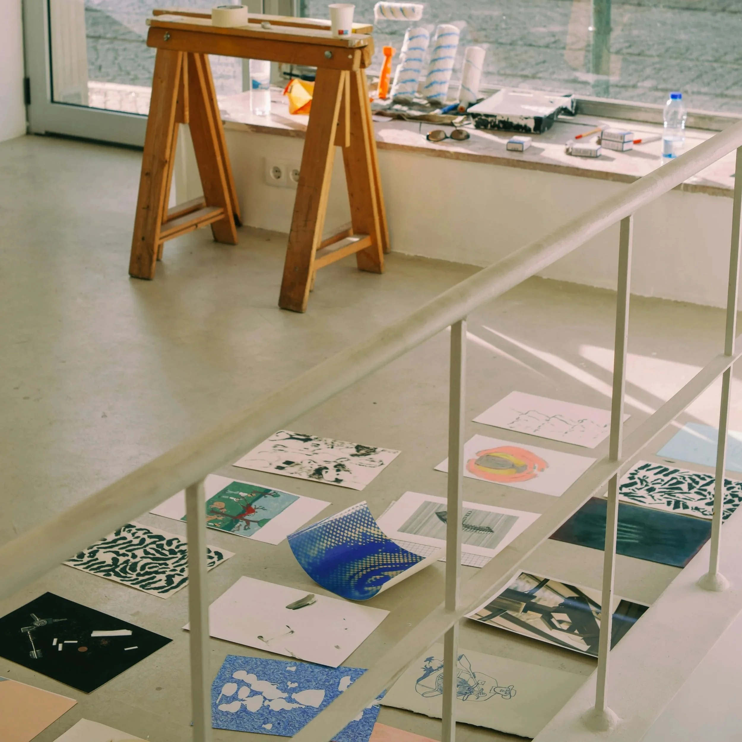 Kunstwerke und Zeichnungen liegen auf dem Boden in einem Raum, im Hintergrund befindet sich ein Holzpult vor einem Fenster.