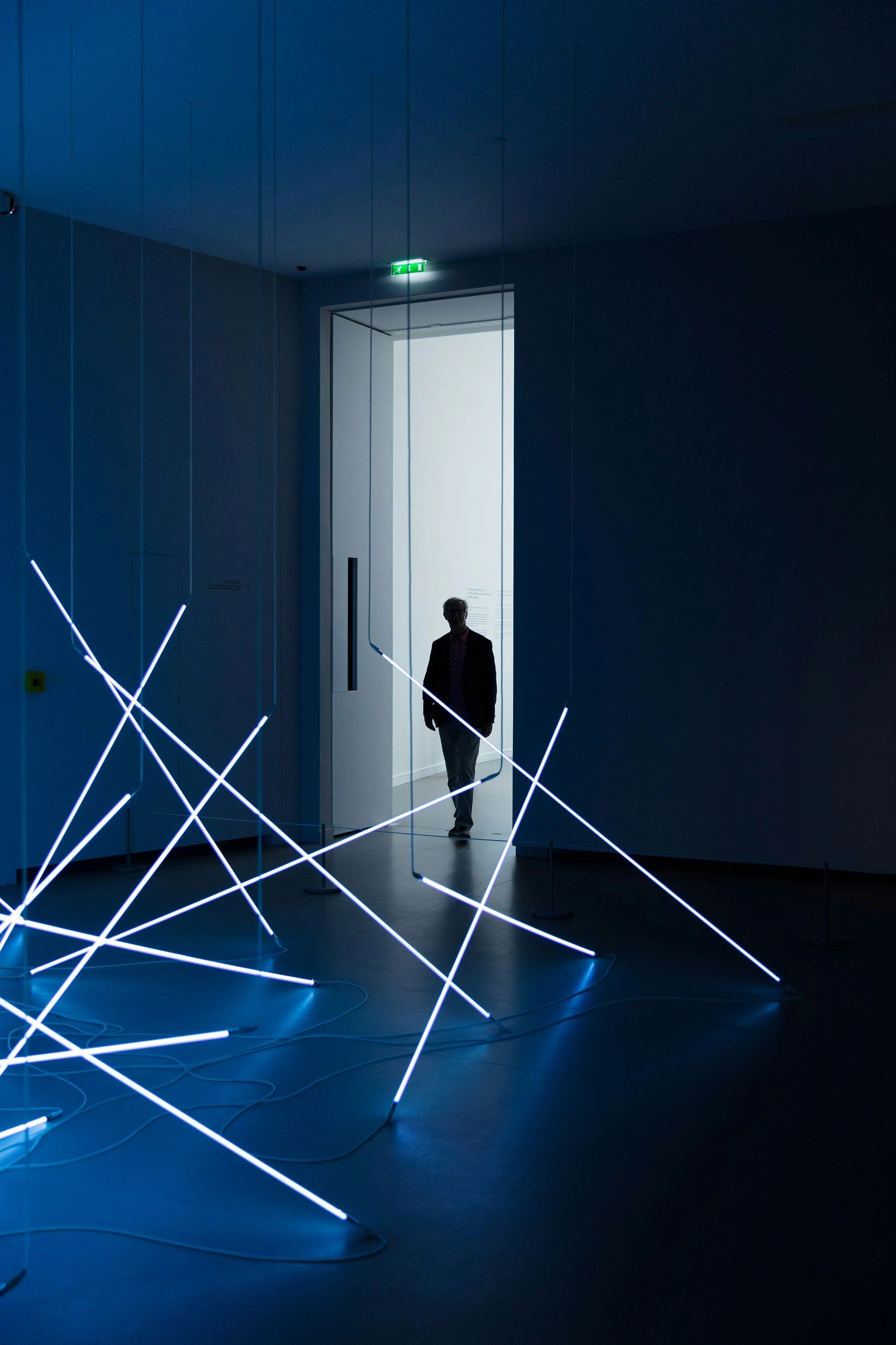 Ein dunkler Raum mit blauer Beleuchtung, in dem ein Mann durch eine offene Tür auf einen hell erleuchteten Raum blickt. Im Raum sind moderne Lichtskulpturen mit leuchtenden weißen Stäben zu sehen.