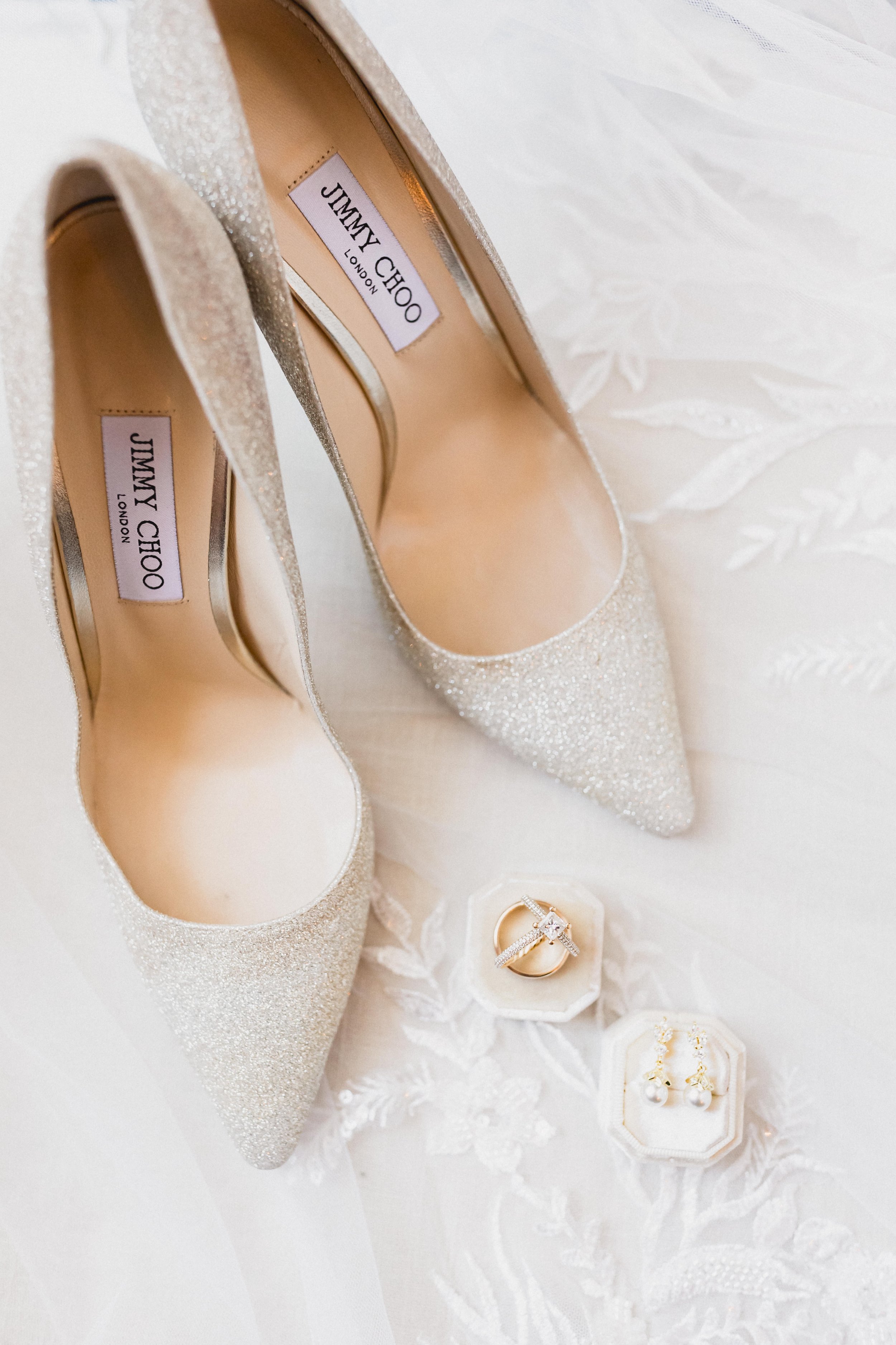A pair of glittering high-heeled shoes, a wedding ring, and pearl earrings on an embroidered white fabric surface.