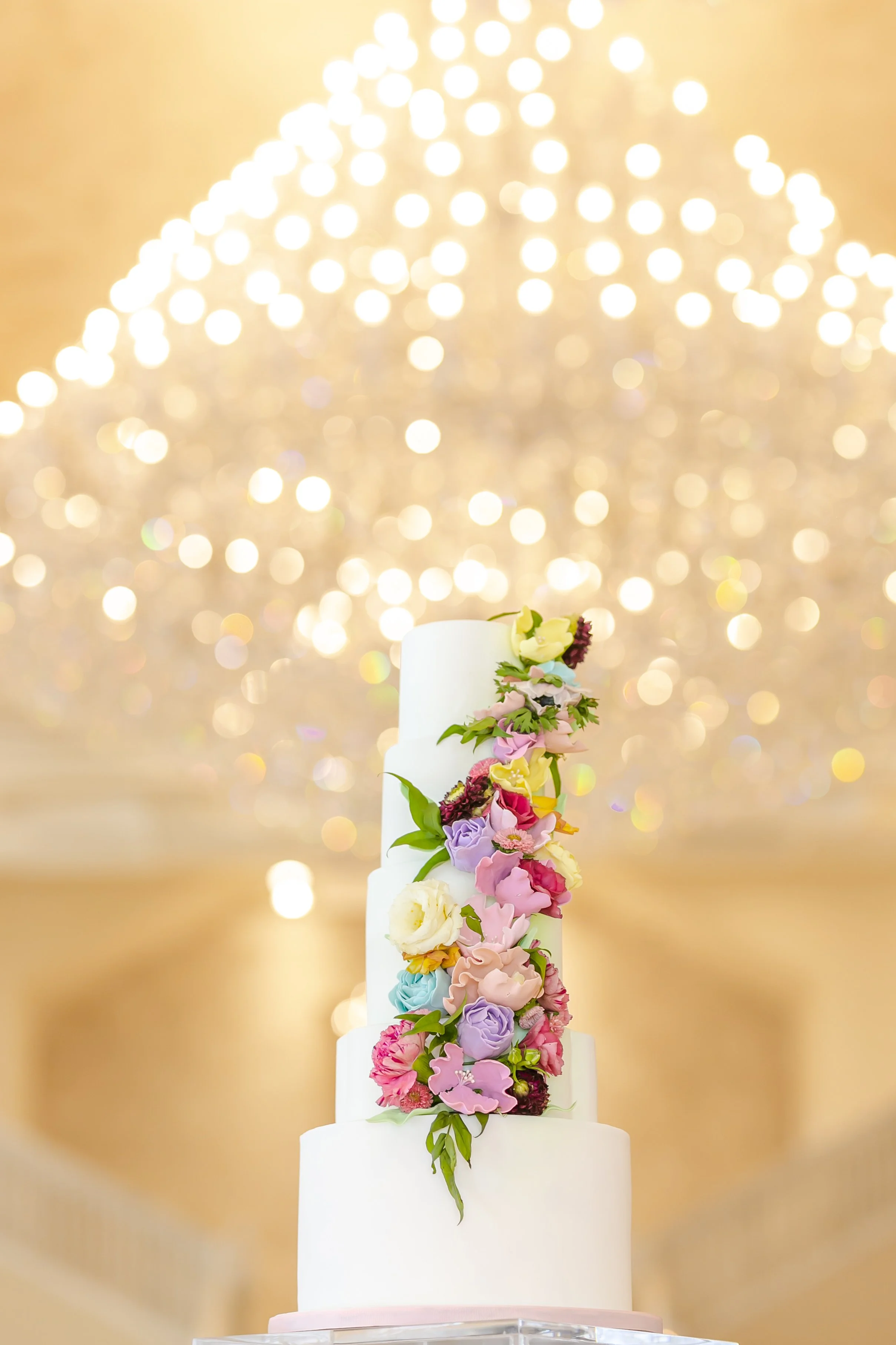 A tall, multi-tiered white wedding cake decorated with colorful flowers, standing against a backdrop of golden, bokeh lights in Atlanta, GA.