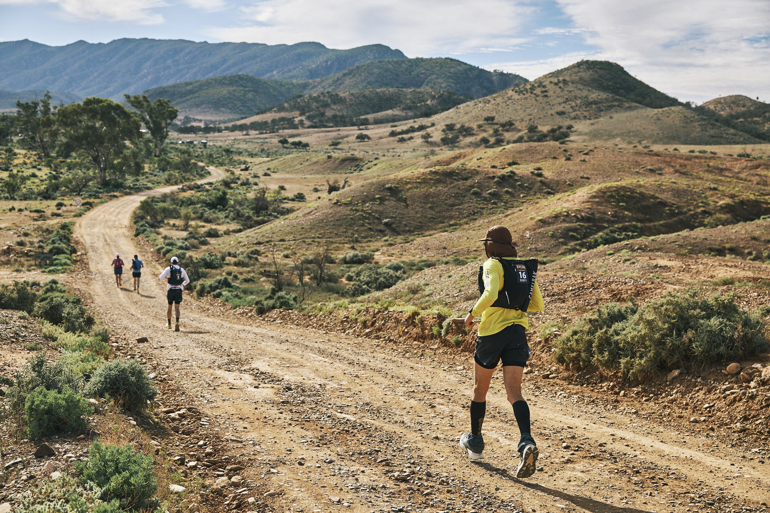A group of four hikers walking on a dirt trail through a mountainous landscape with hills and sparse vegetation.