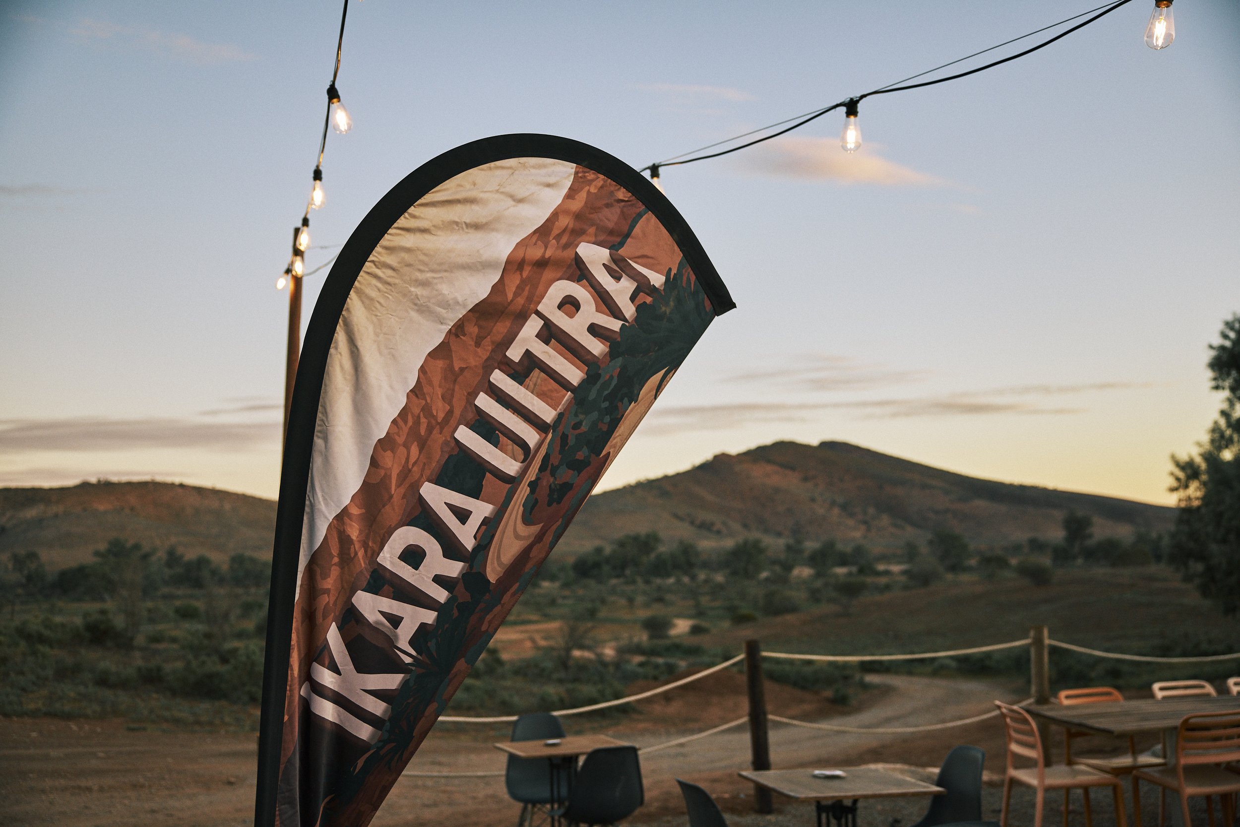 A colorful banner with the word "Outback" hangs outdoors during sunset, with string lights overhead and hills in the background.