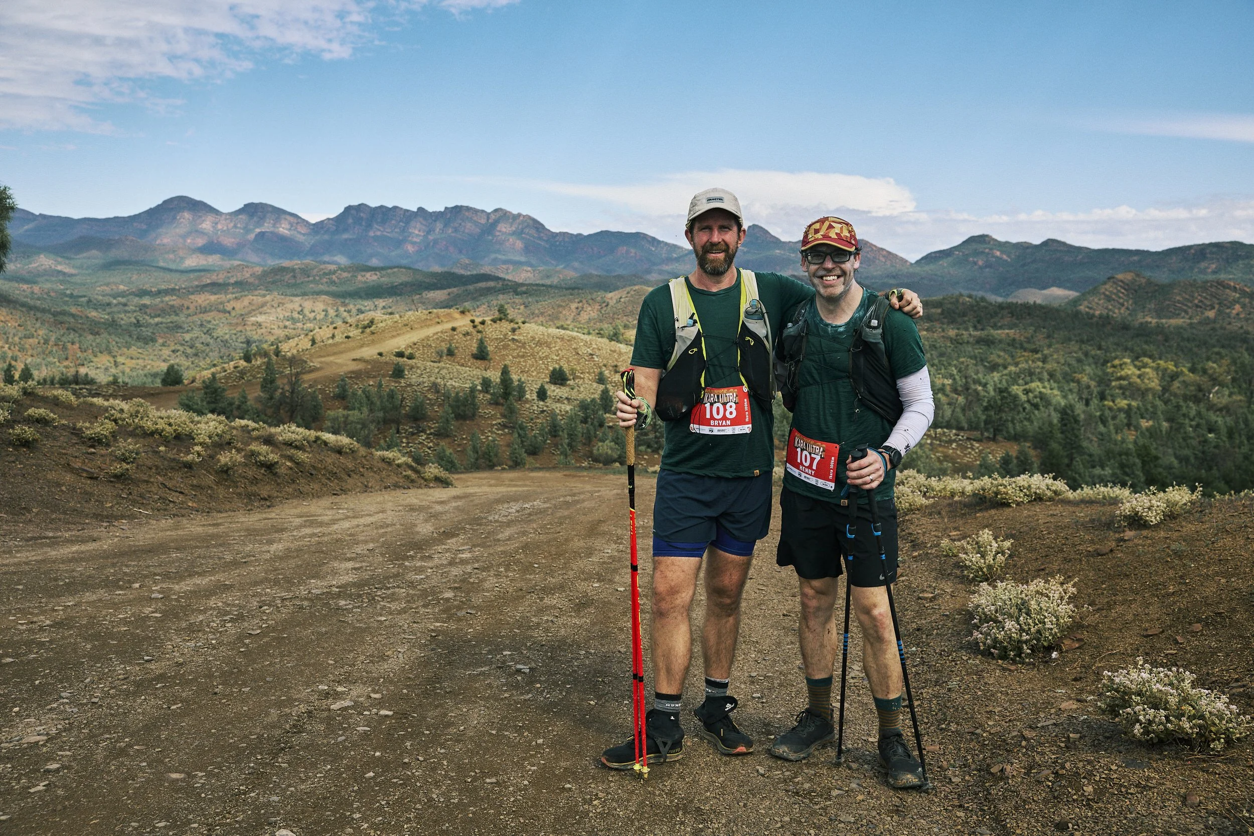 Two men standing outdoors in a scenic mountainous landscape with blue sky, wearing athletic clothing and holding trekking poles. They are smiling, wearing race bibs, and appear to be participating in a trail running event.