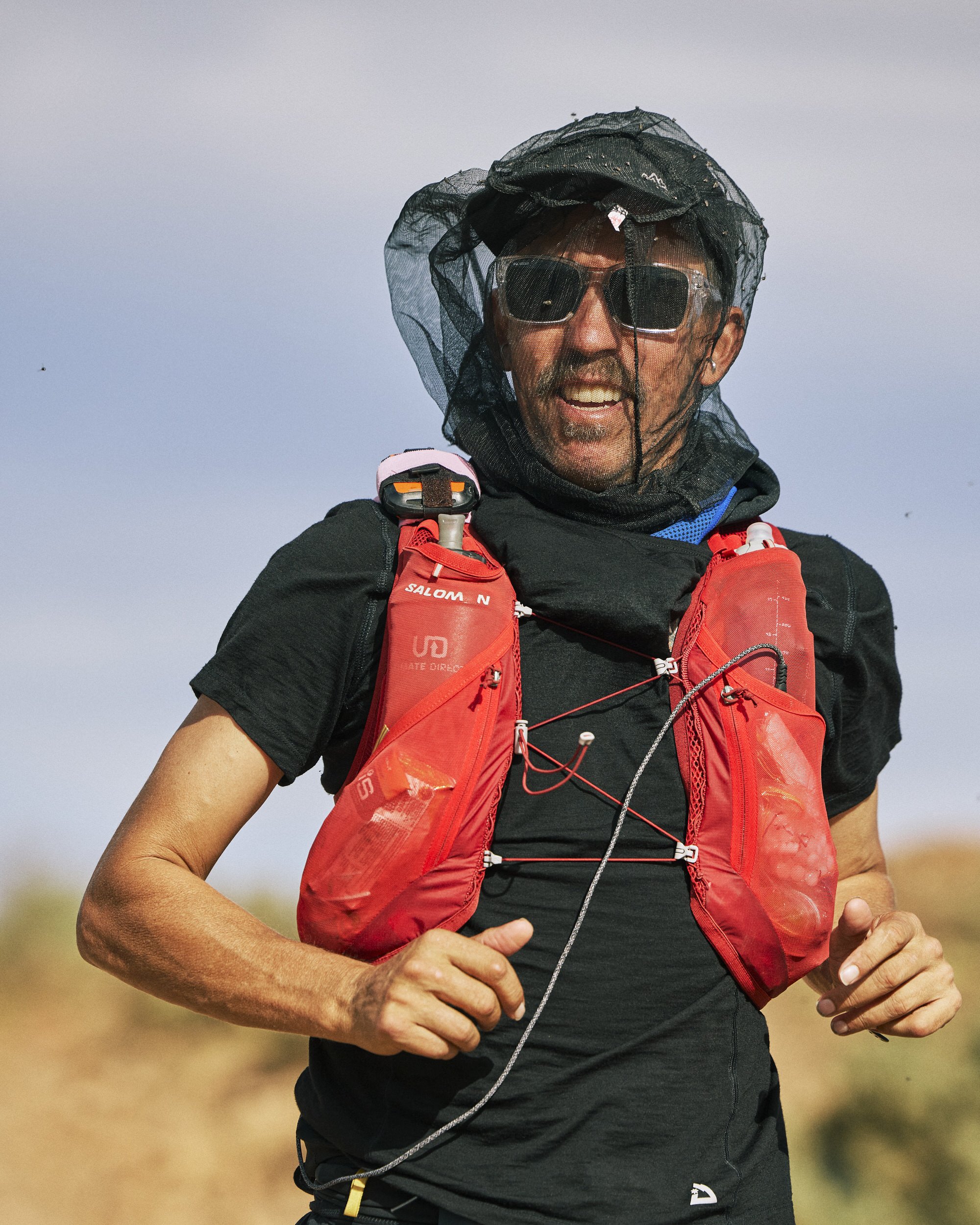 A man trail running outdoors, wearing sunglasses, a black shirt, and a red hydration vest, with a black sun hat and a black netted head covering. He is smiling and holding a running device in his left hand, with a blurry background of grass and sky.