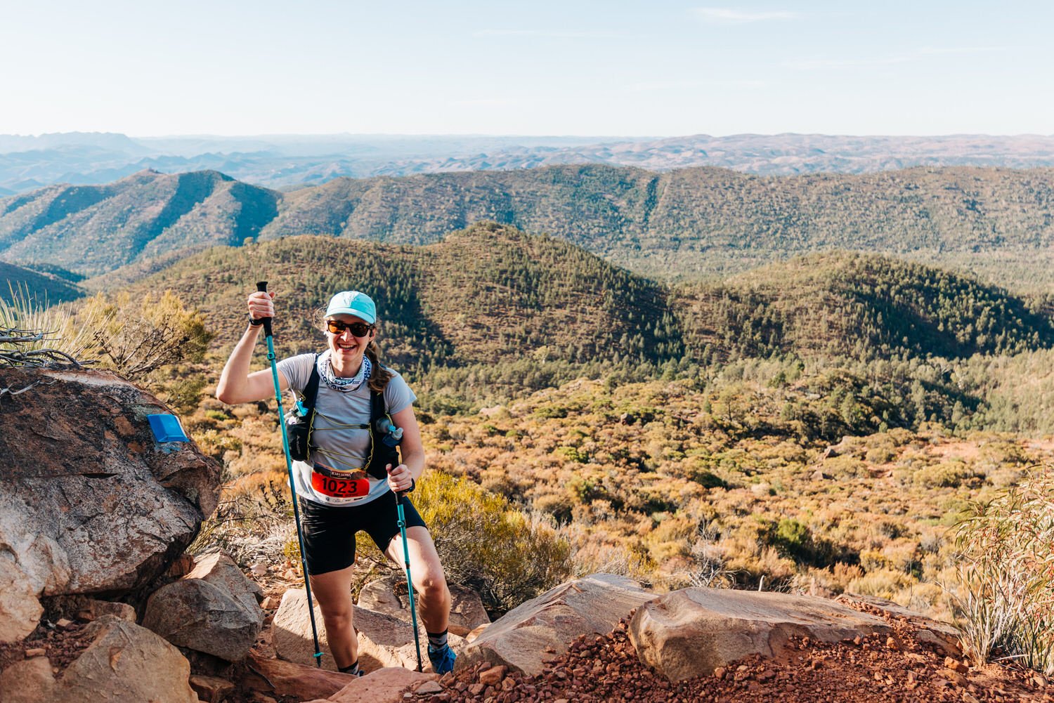 A woman trail running on a rocky path with mountains and a forest in the background, wearing a blue cap, sunglasses, a gray shirt, black shorts, and a race bib.