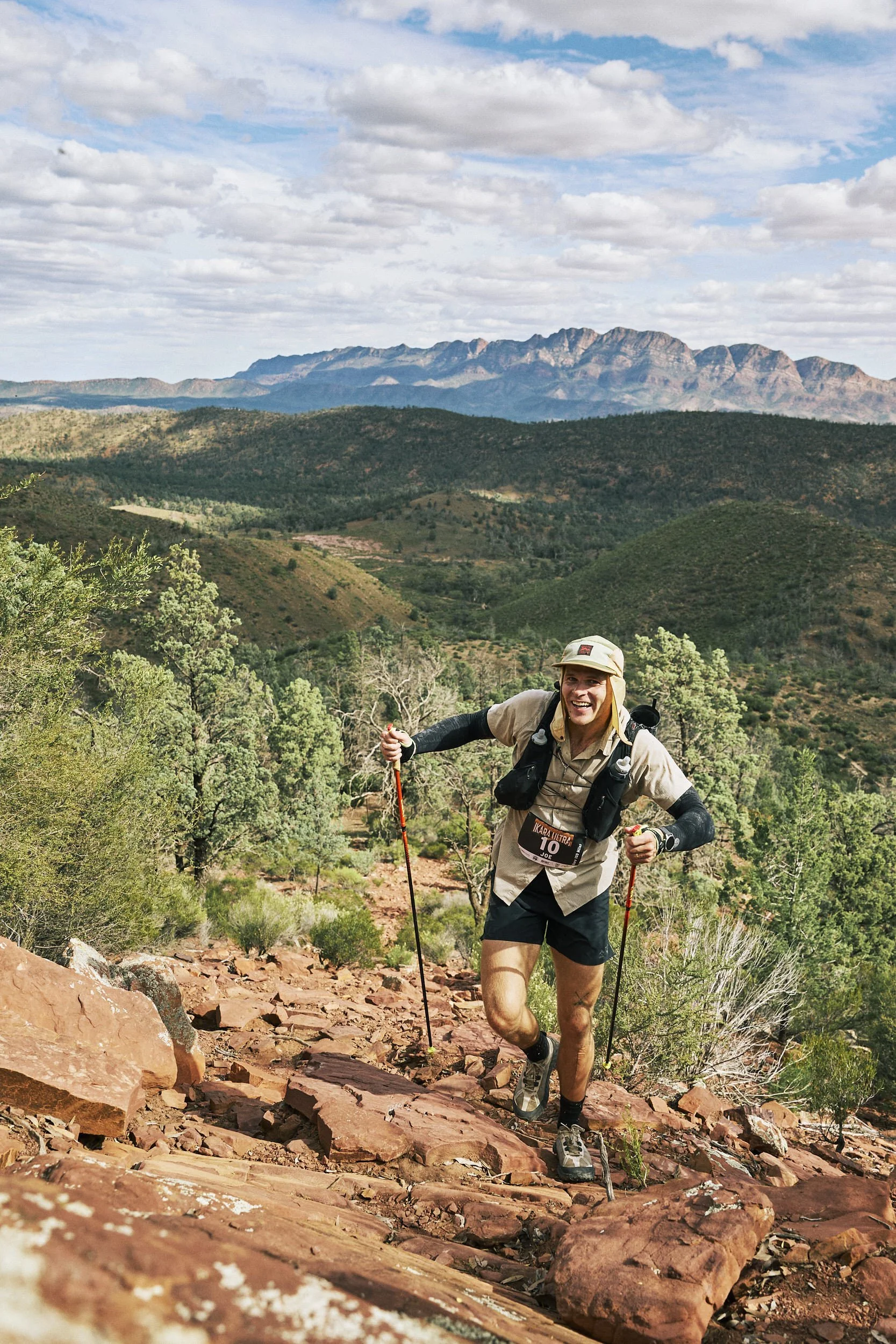 Person running on a dirt trail in a mountainous area, wearing a yellow jacket, black shorts, and a black backpack.