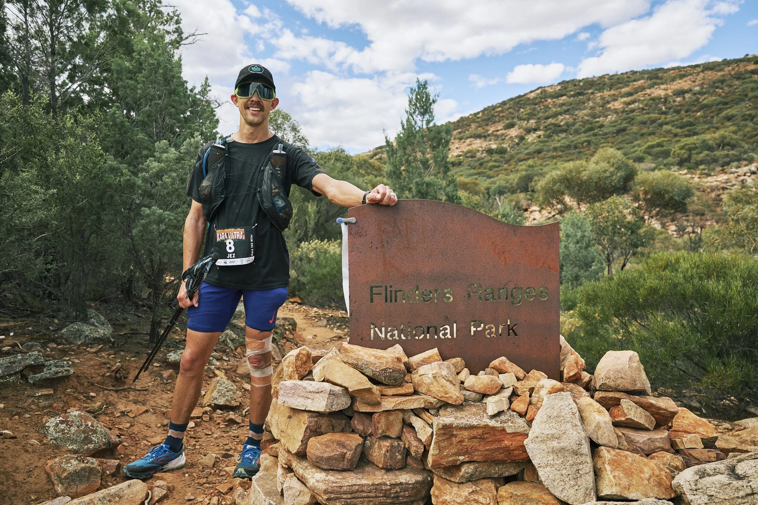 A man wearing a black cap, sunglasses, a black athletic shirt, blue shorts, and running shoes, standing next to a sign that reads 'Flinders Ranges National Park' surrounded by rocks, with green trees and a hill in the background under a partly cloudy