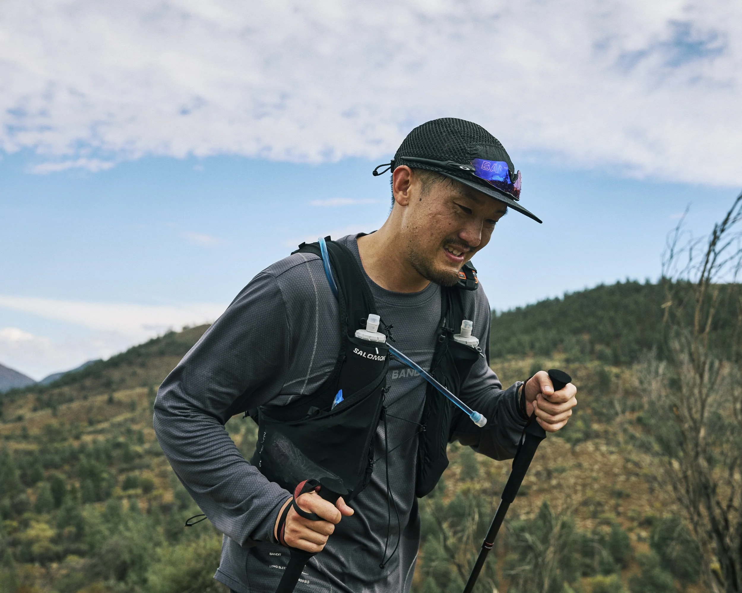 A man wearing a black cap with purple sunglasses on the brim, a gray hiking shirt, and a black backpack is outdoors on a trail, holding a hiking pole, with mountainous terrain and a partly cloudy sky in the background.