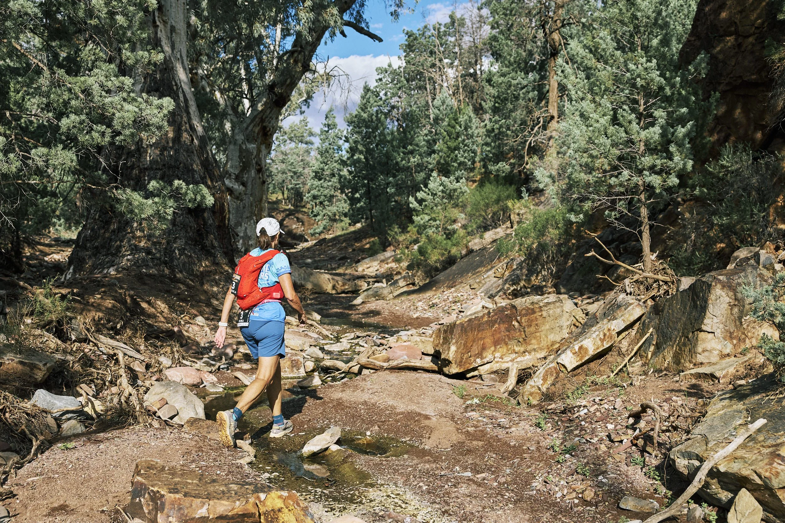A person hiking in a forested area with trees and rocks, wearing a red backpack, blue shorts, a white cap, and hiking shoes.