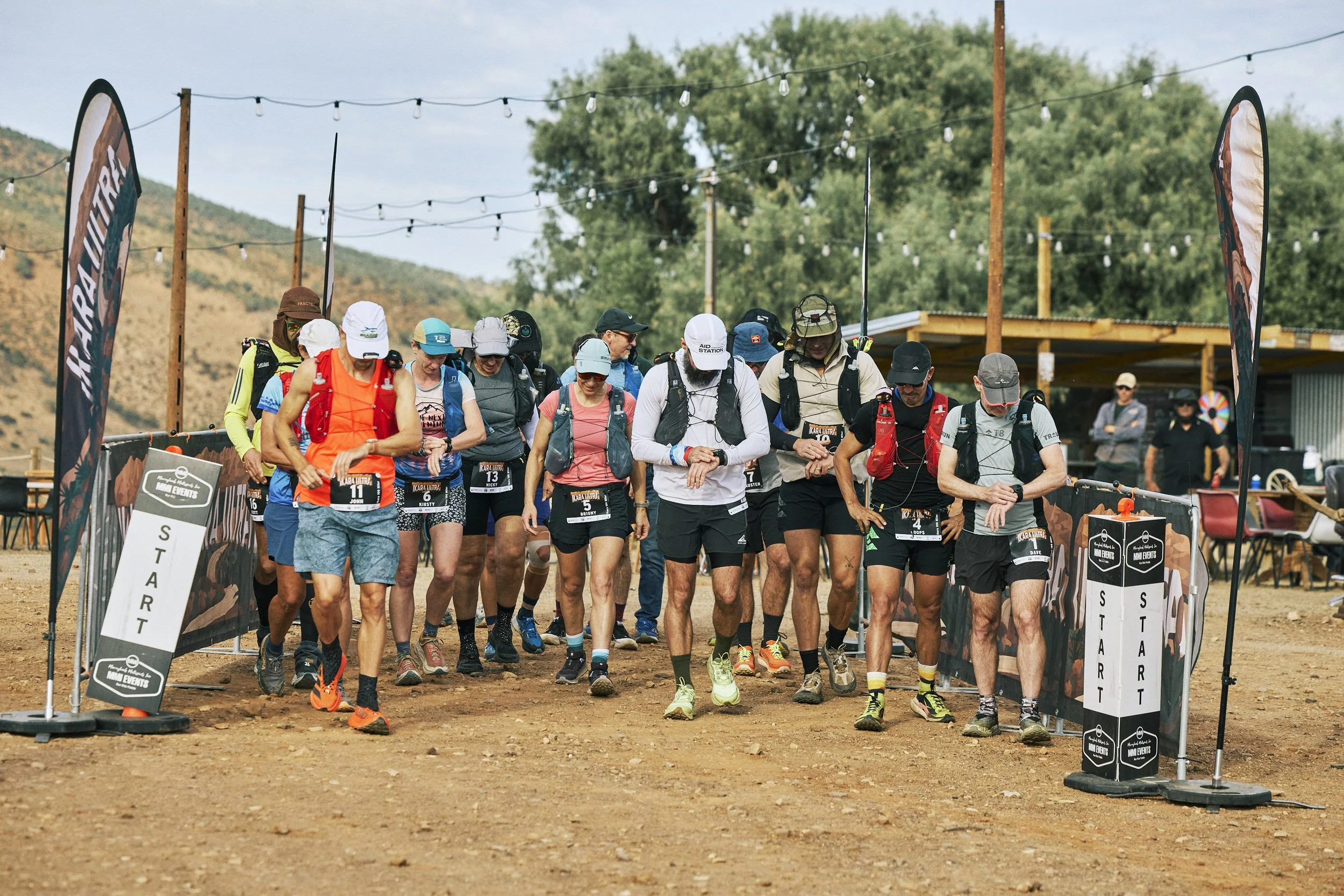 Group of runners at the starting line of an outdoor race event, with race signs and flags, in a rural setting with hills and trees in the background.