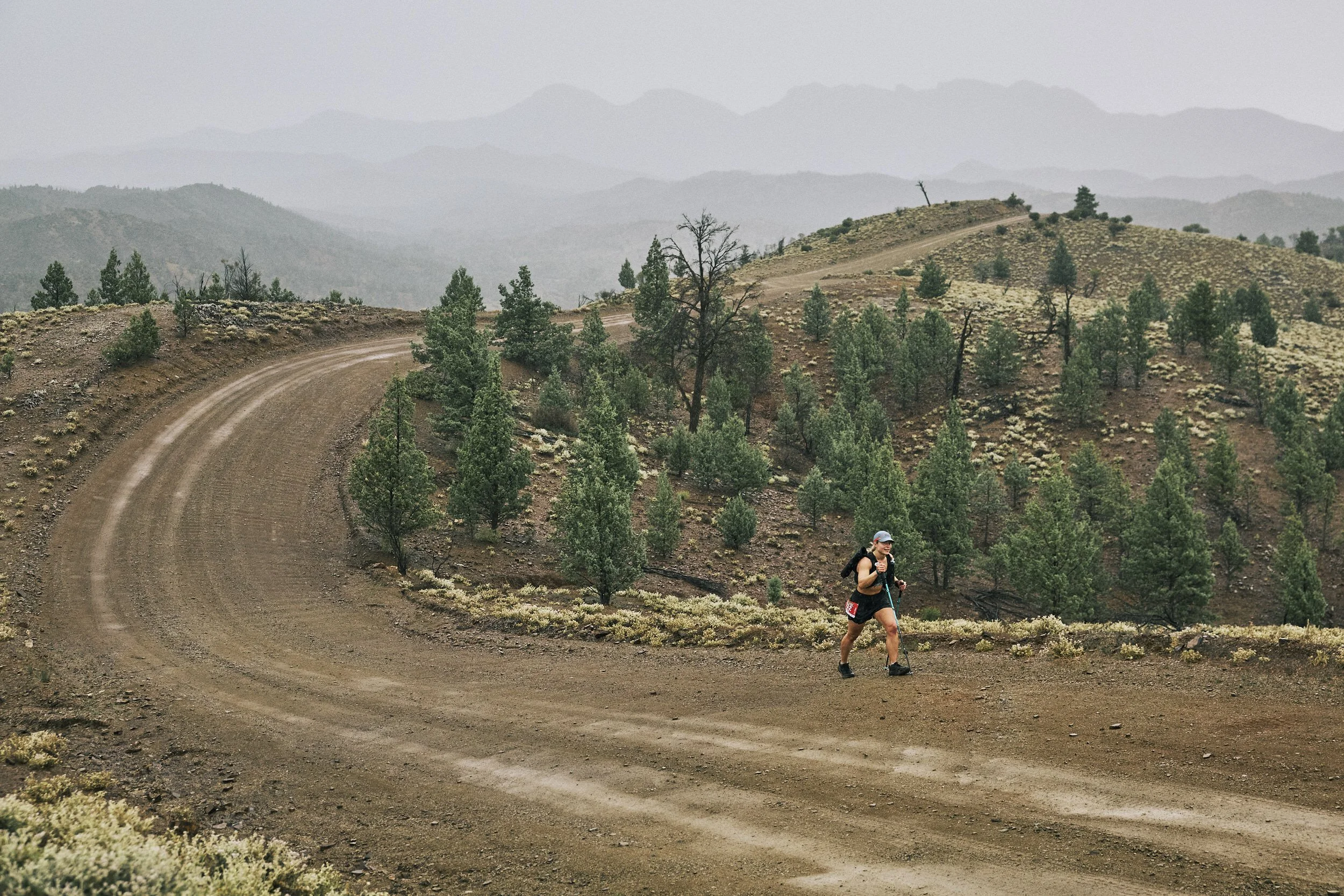 A woman running along a winding dirt trail in a hilly, semi-arid landscape with scattered trees and distant mountain ranges under an overcast sky.