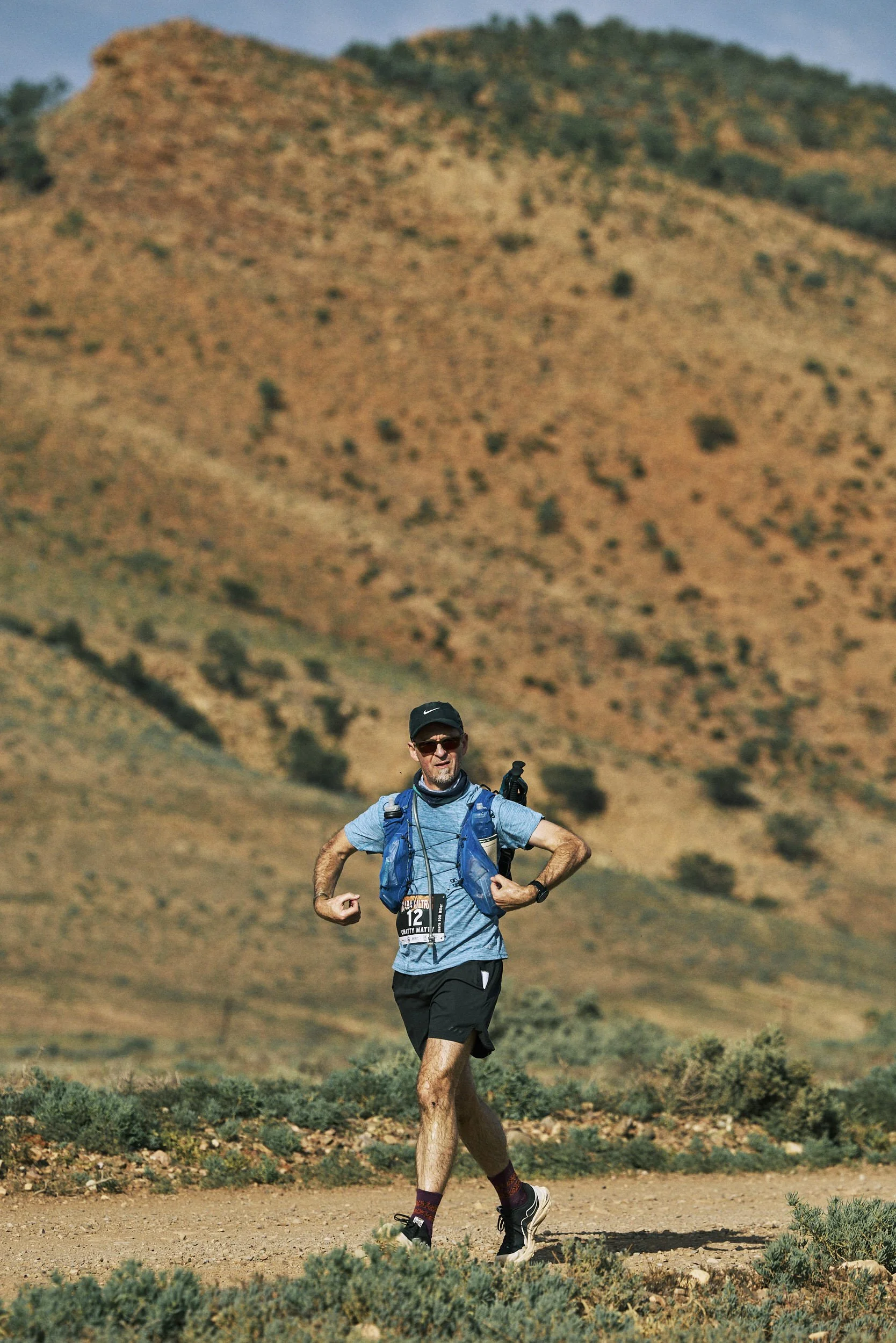 A man in athletic gear running on a trail in a dry, hilly landscape with sparse vegetation.