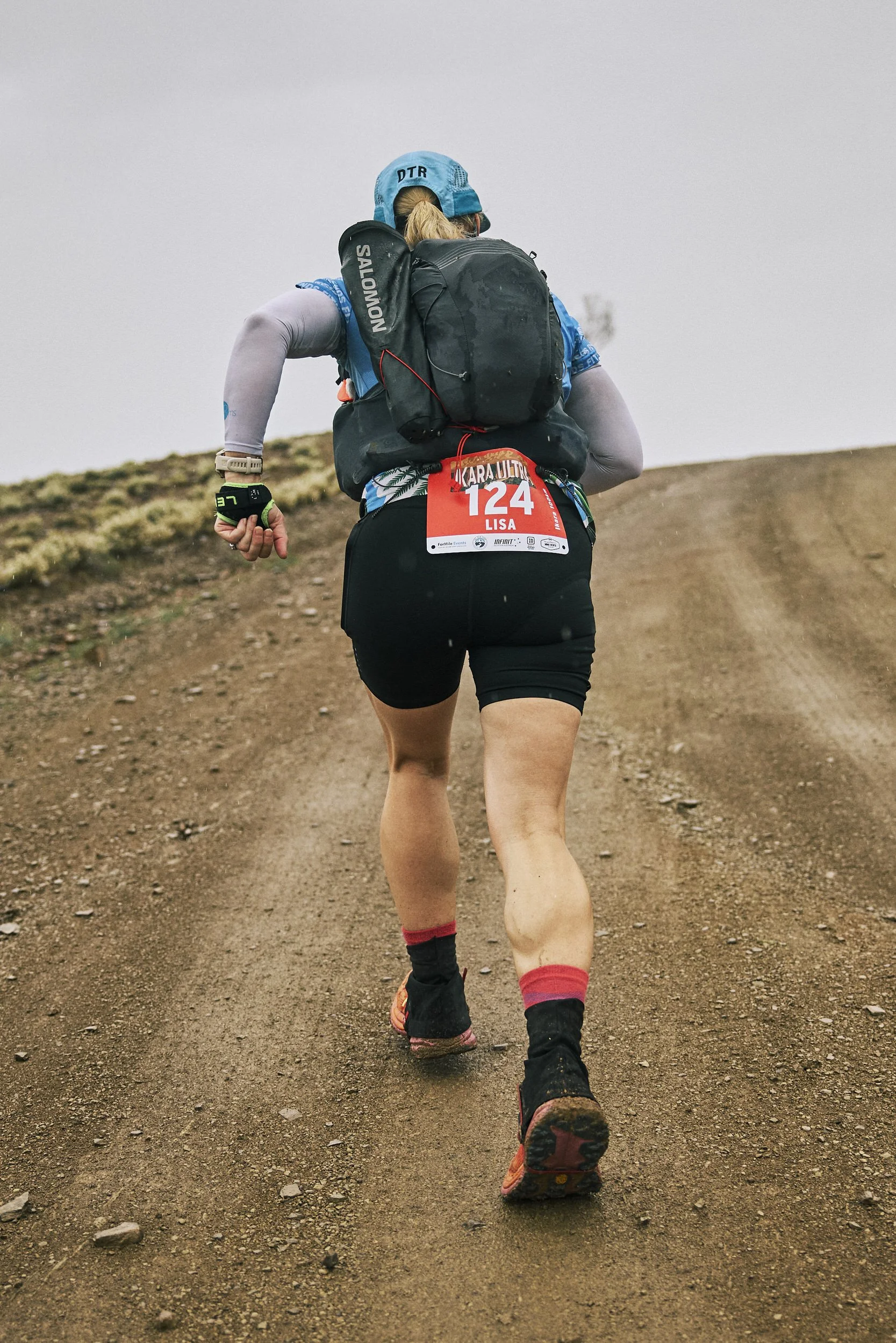 A female trail runner in athletic clothing and gear running on a dirt trail in an outdoor setting with a cloudy sky.