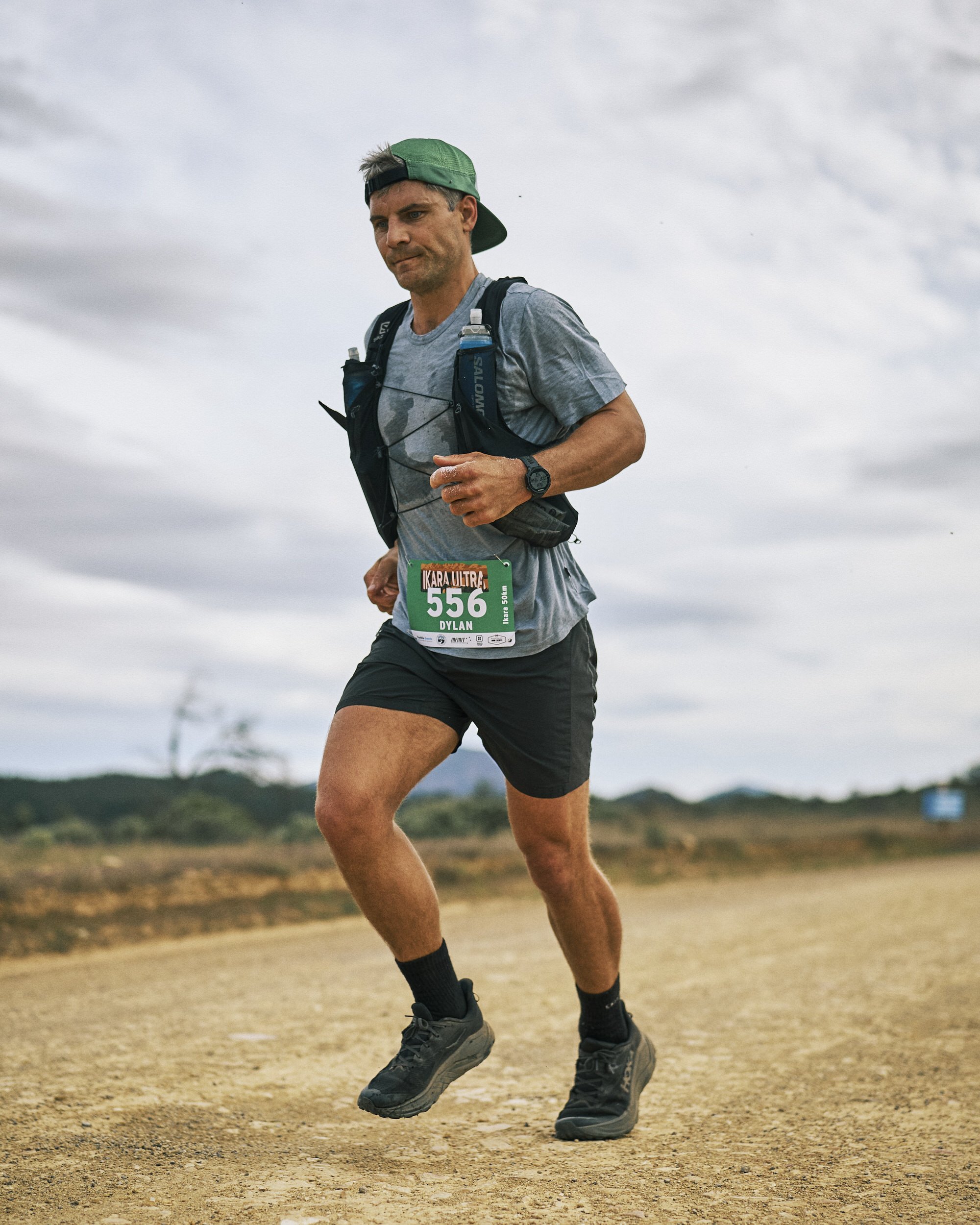Male runner in athletic gear running on country dirt road under cloudy sky, wearing a green cap backwards, gray T-shirt, black shorts, and hiking shoes.