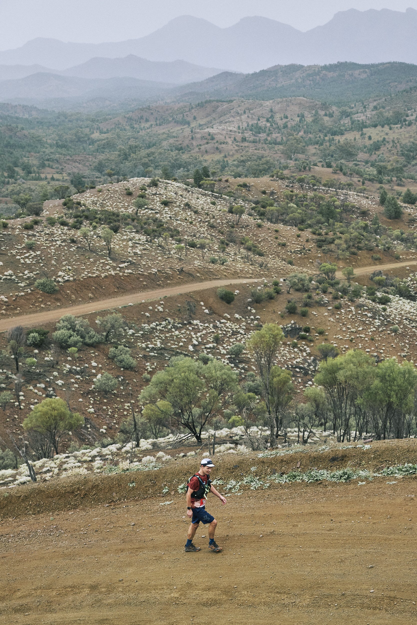 A person trail running on a dirt path through a hilly landscape with sparse trees, with rolling hills and mountains in the background.