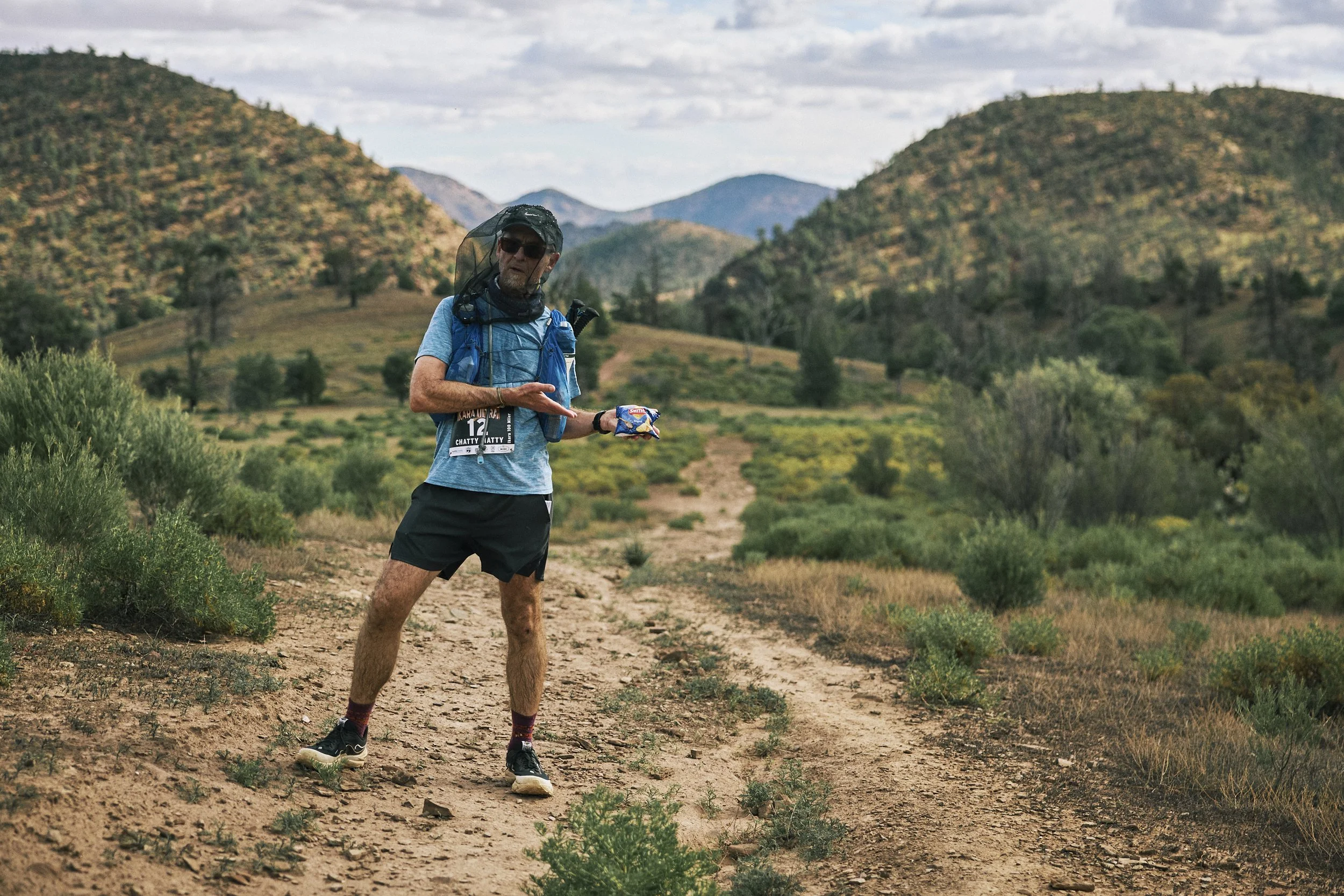 A man running on a dirt trail in a hilly, desert landscape during daytime, with mountains and sparse vegetation in the background.