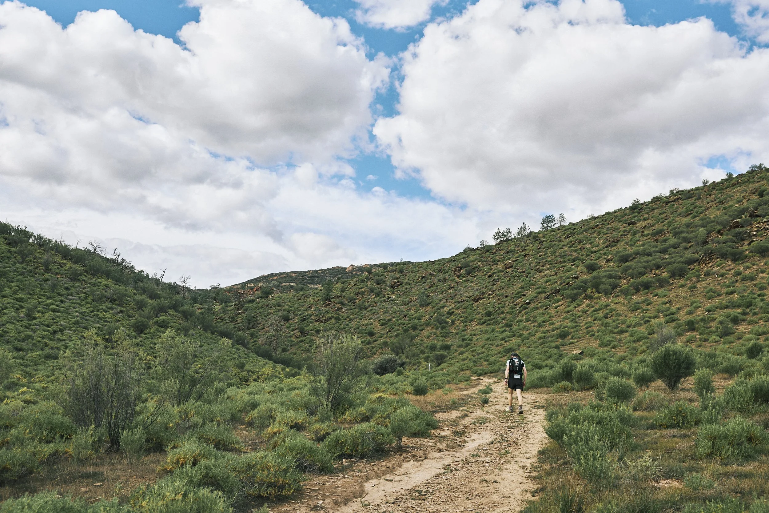 A person hiking on a dirt trail through green hills with a partly cloudy sky overhead.