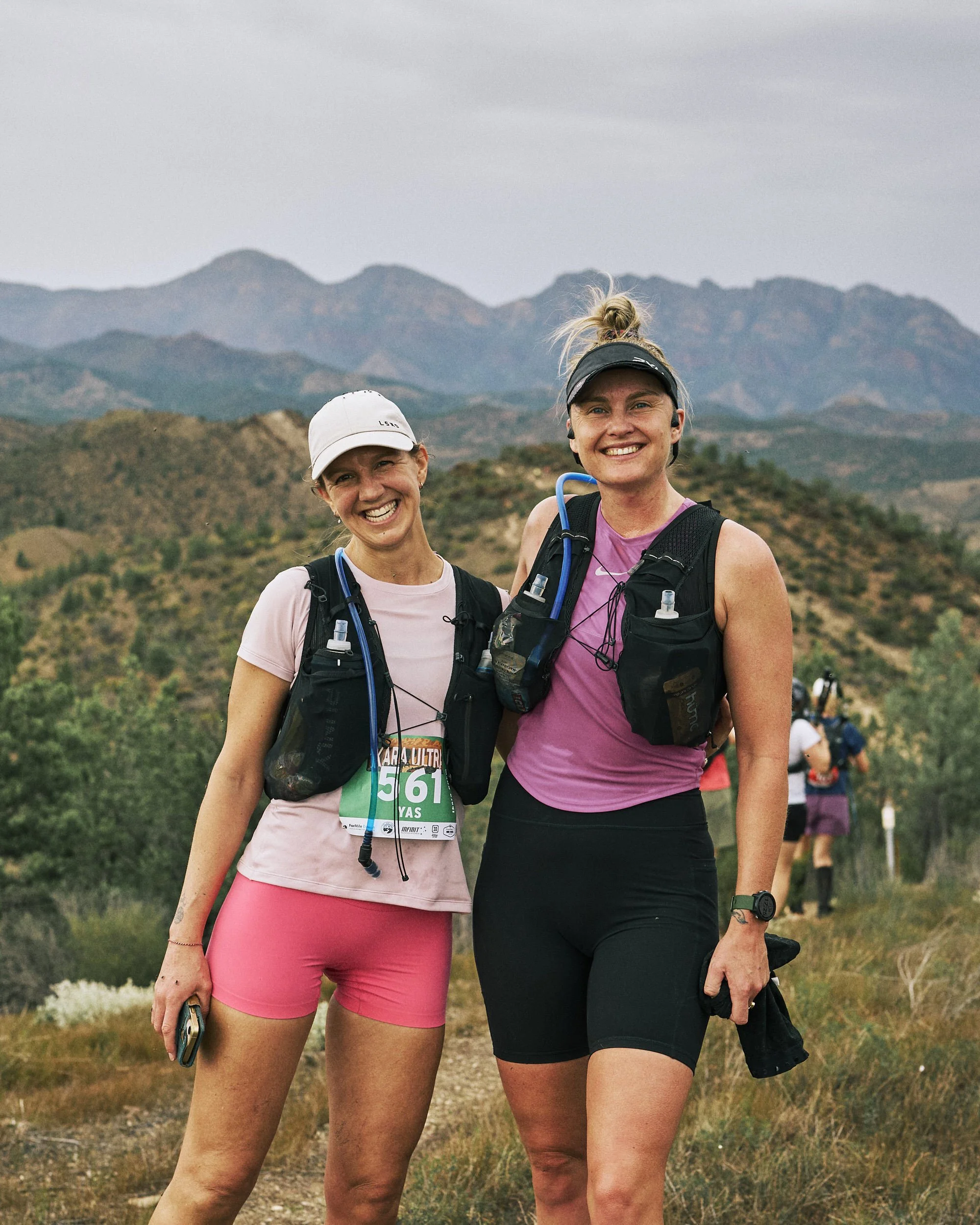Two women standing outdoors in a mountainous area, smiling at the camera. They are dressed in athletic gear and carrying hydration packs, indicating they are participating in a trail or mountain race.