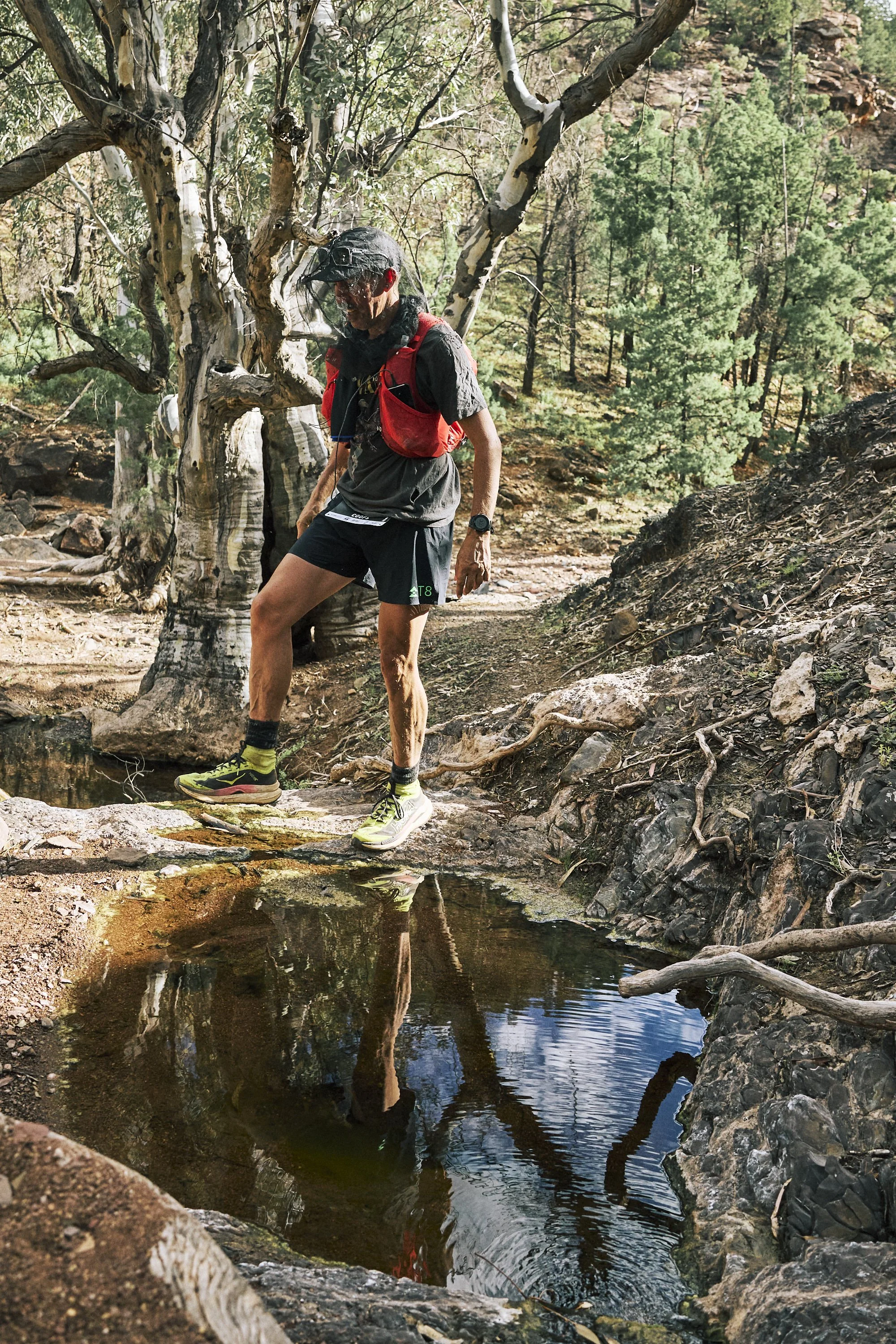 A trail runner with a red vest and black shorts crossing a small stream on a dirt trail in a wooded area with pine trees and rocky terrain.