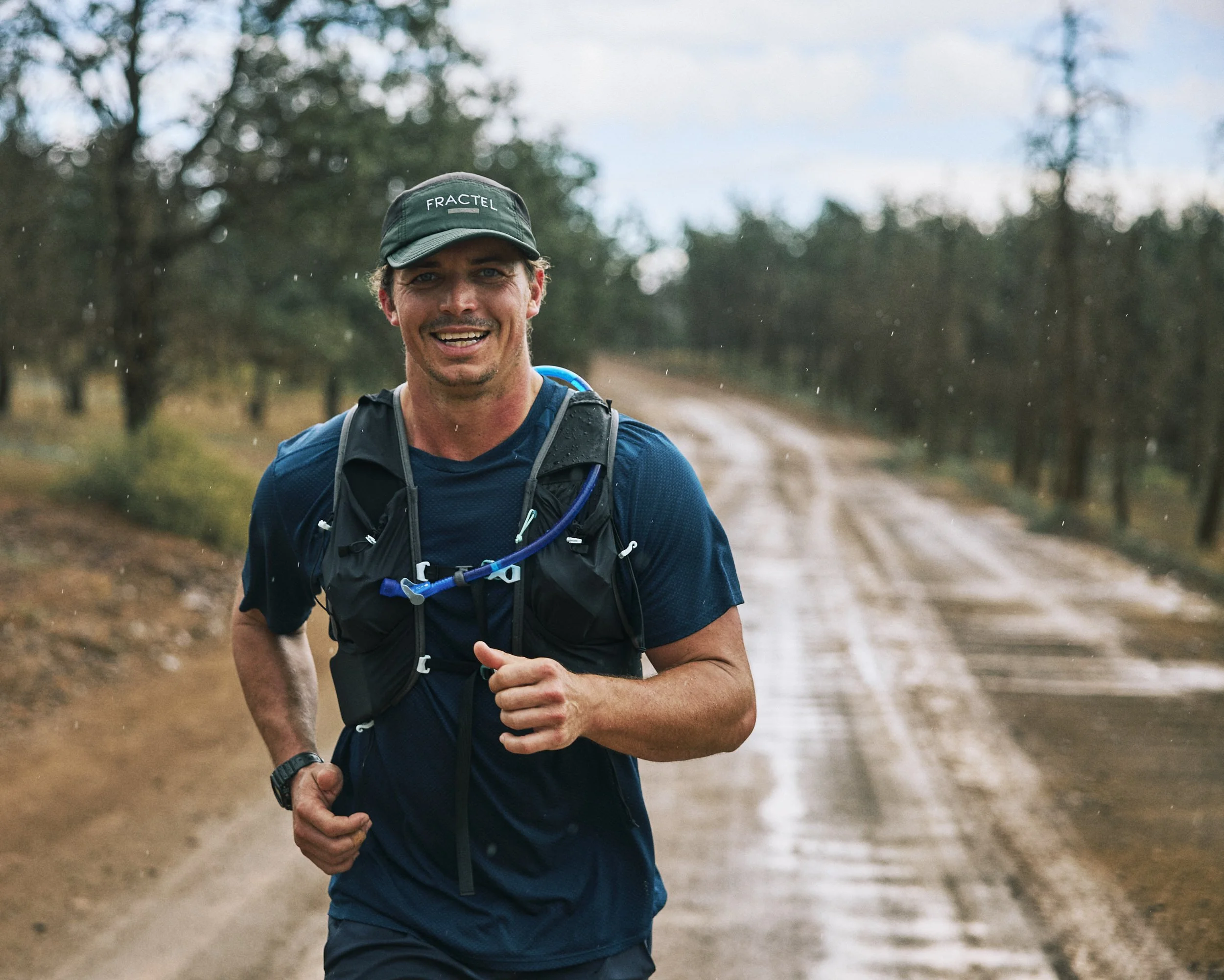 A man smiling and running outdoors on a dirt trail, wearing a black cap with the word 'FRACTEL', a blue shirt, and a backpack.