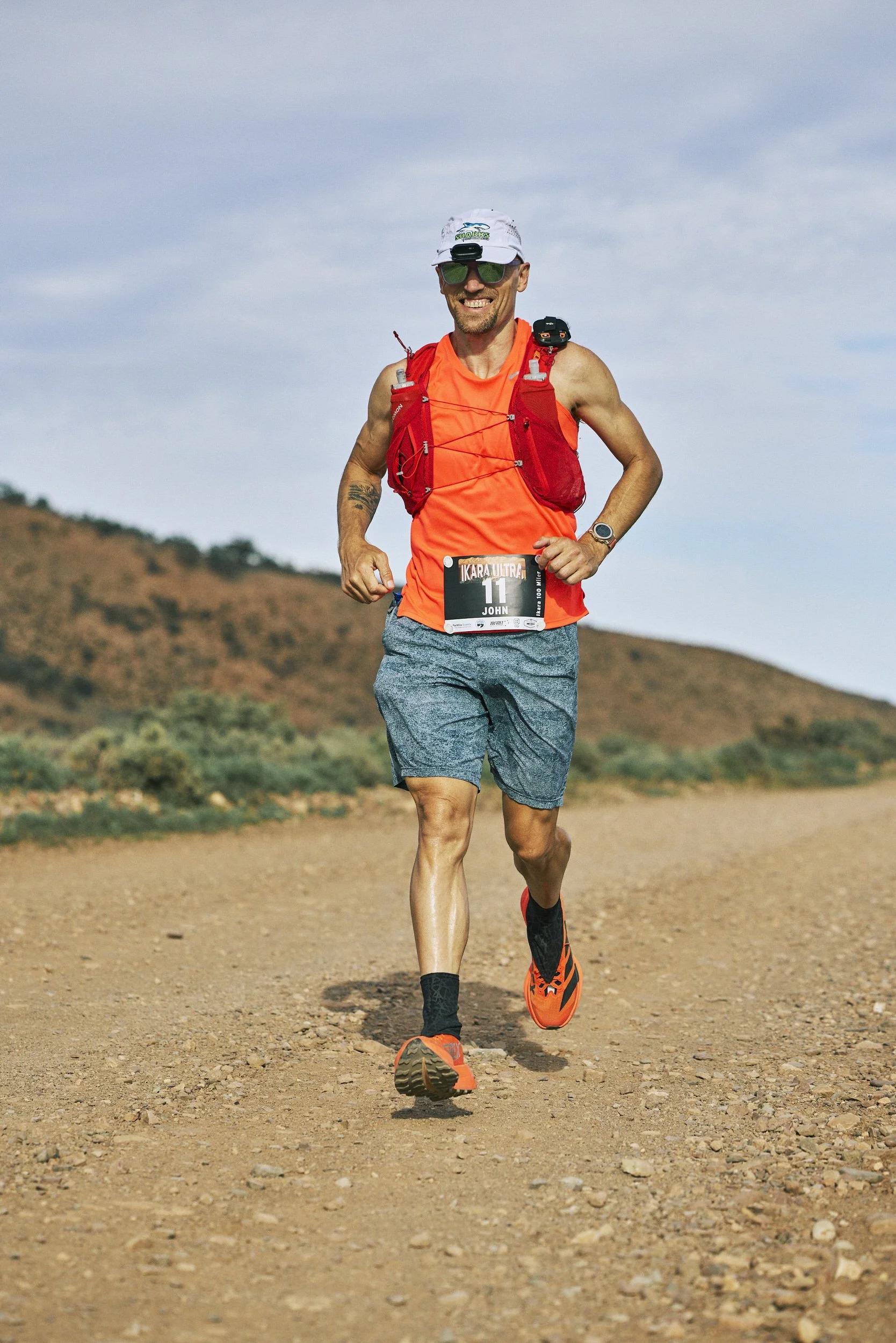 A man running outdoors on a dirt trail. He is wearing a white cap, sunglasses, a bright orange sleeveless shirt, gray shorts, and bright orange running shoes. He has a red hydration vest and a race bib with the number 11. The background features a la