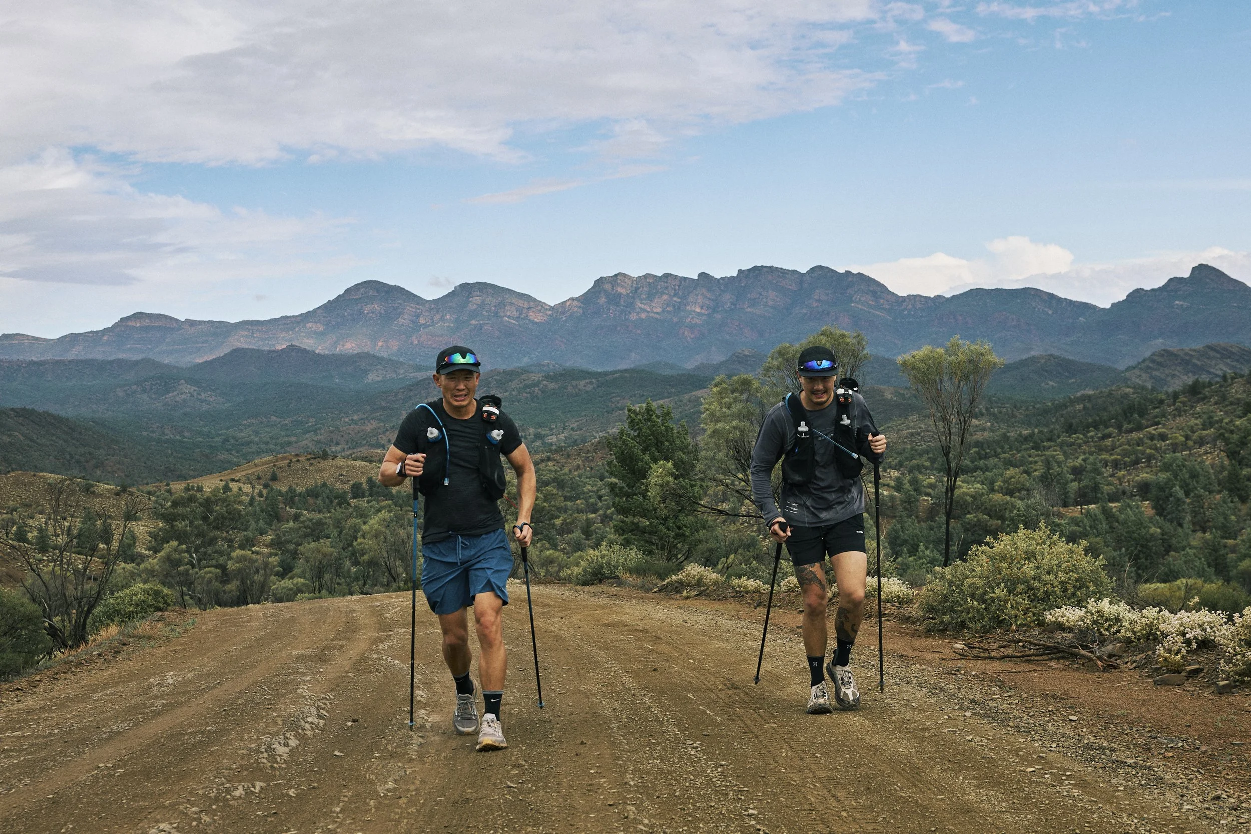 Two people hiking on a dirt trail in a mountainous landscape with trees and a blue sky.