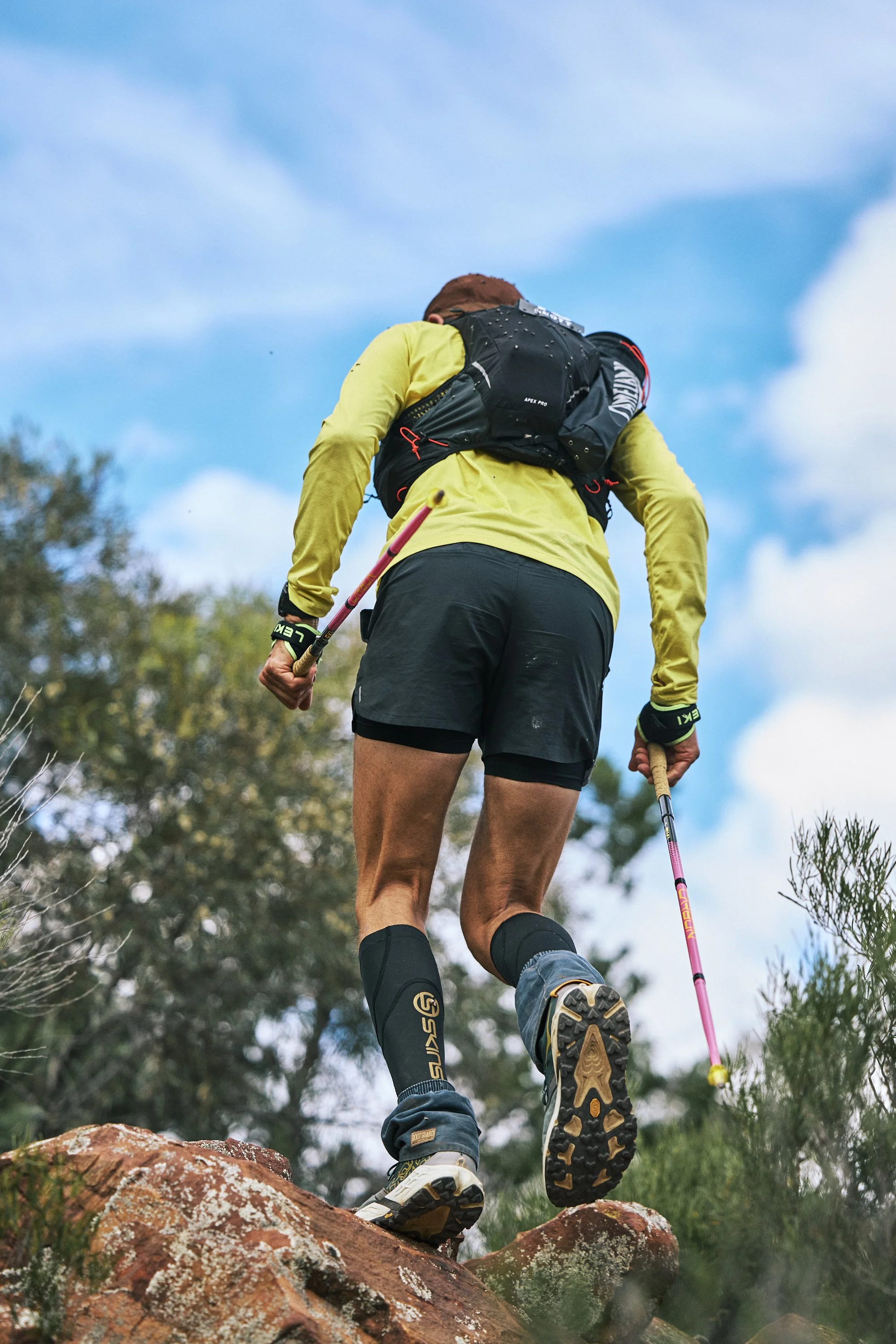 A person hiking outdoors, climbing over rocks with trekking poles, wearing a yellow long-sleeve shirt, black shorts, knee-high black socks, and a black backpack, against a background of blue sky and trees.