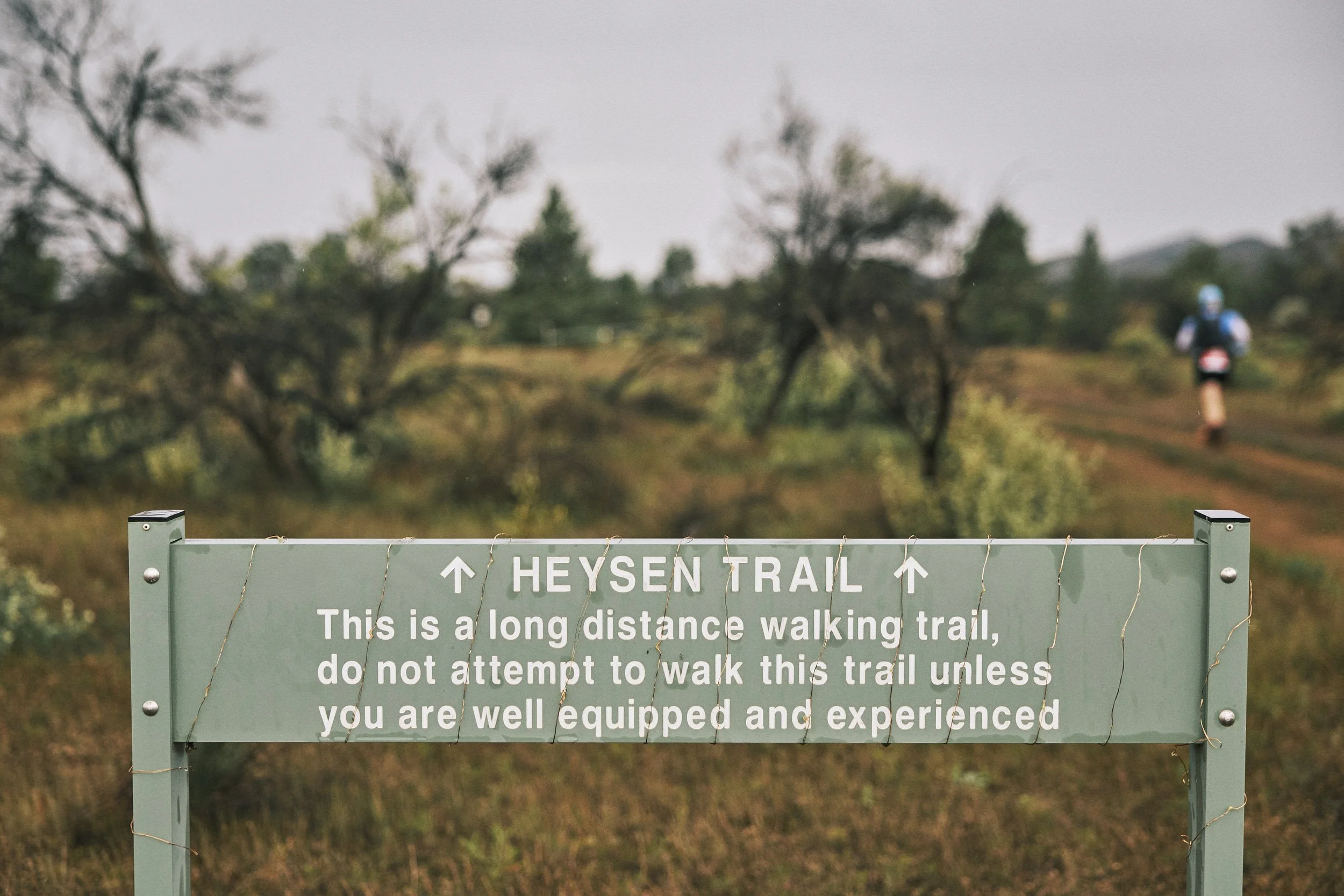 A sign at the entrance of Heysen Trail reading, "This is a long distance walking trail. Do not attempt to walk this trail unless you are well equipped and experienced." The background shows blurred trees and a hiker in outdoor gear walking along the 