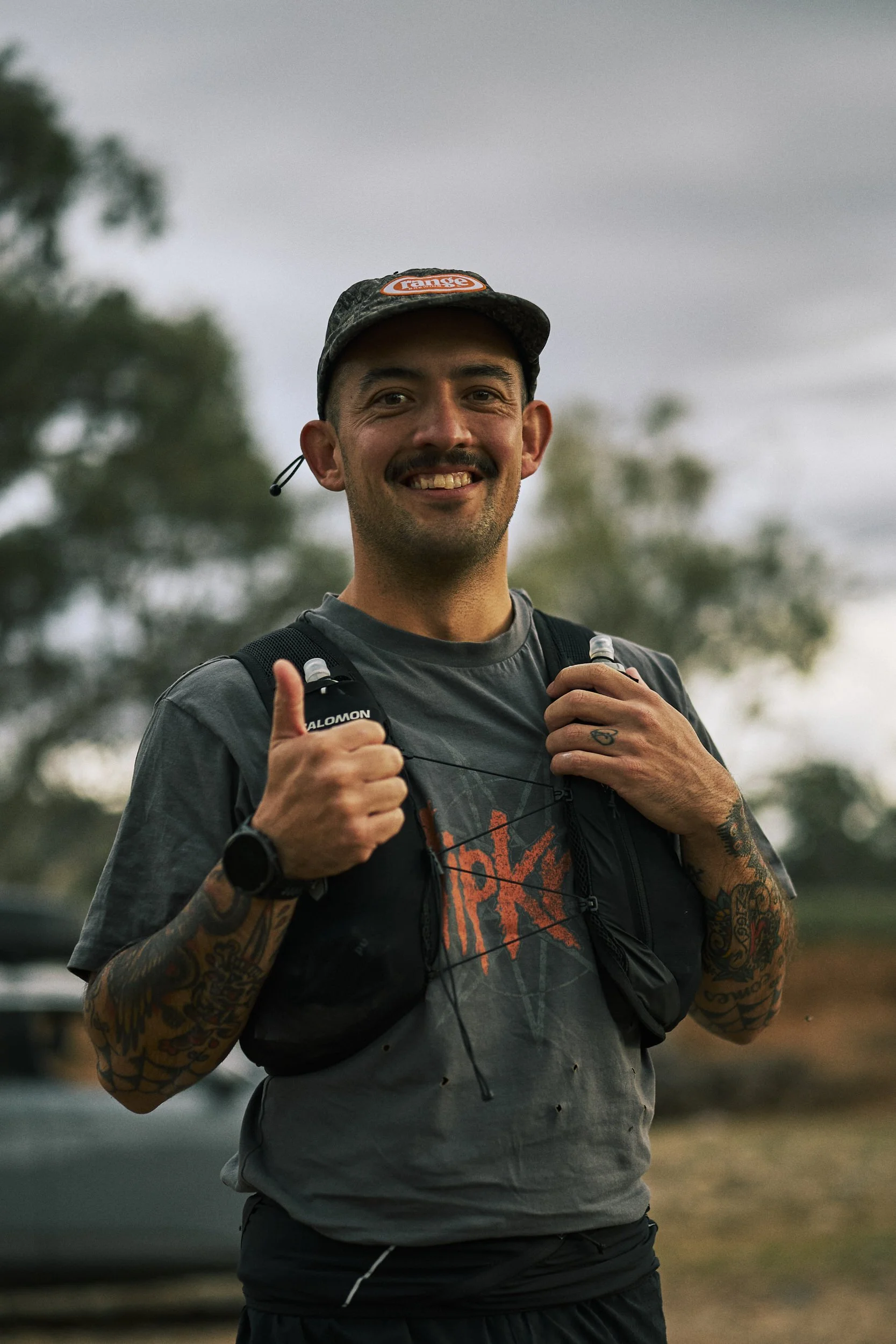 Male trail runner smiling and giving a thumbs-up while holding his hydration pack straps, outdoors with cloudy sky and trees in the background.