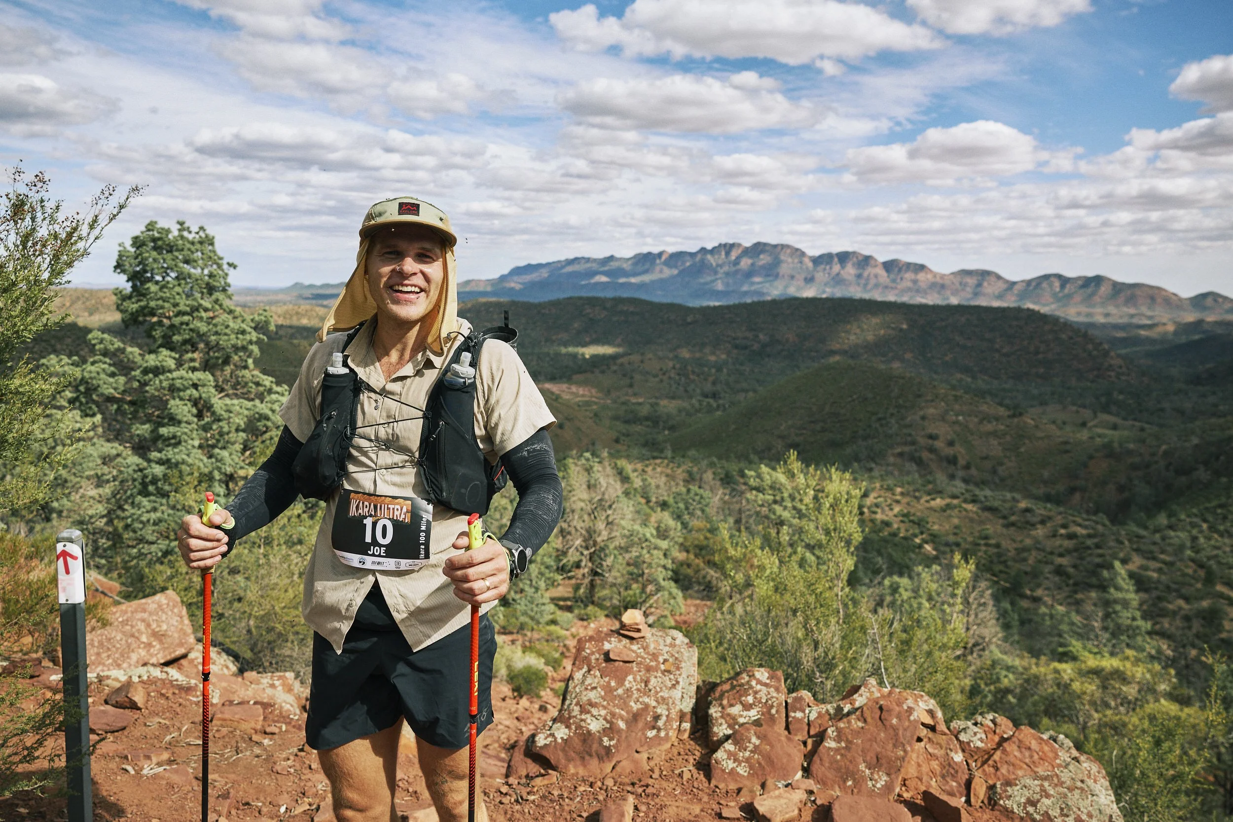 A man smiling while running on a trail in a mountainous area during daytime, wearing sporty outdoor clothing, a hat, and using trekking poles.