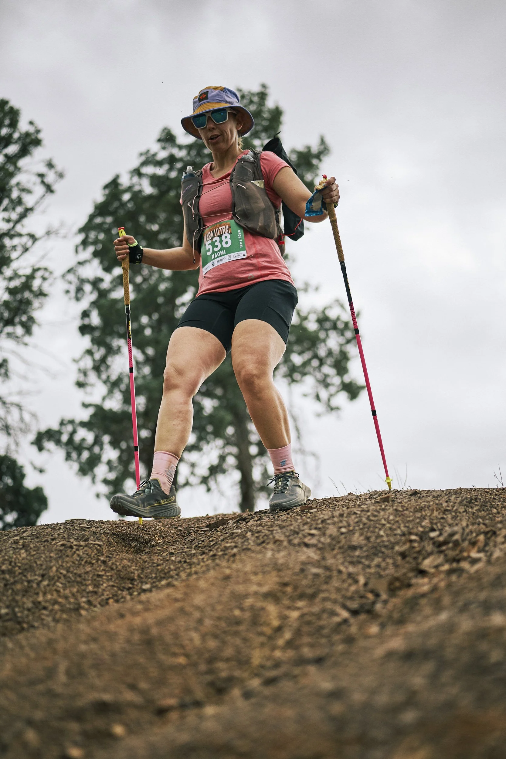 A woman trail running outdoors on a dirt path with trees in the background, wearing a hat, sunglasses, a pink shirt, black shorts, hiking shoes, and carrying trekking poles.