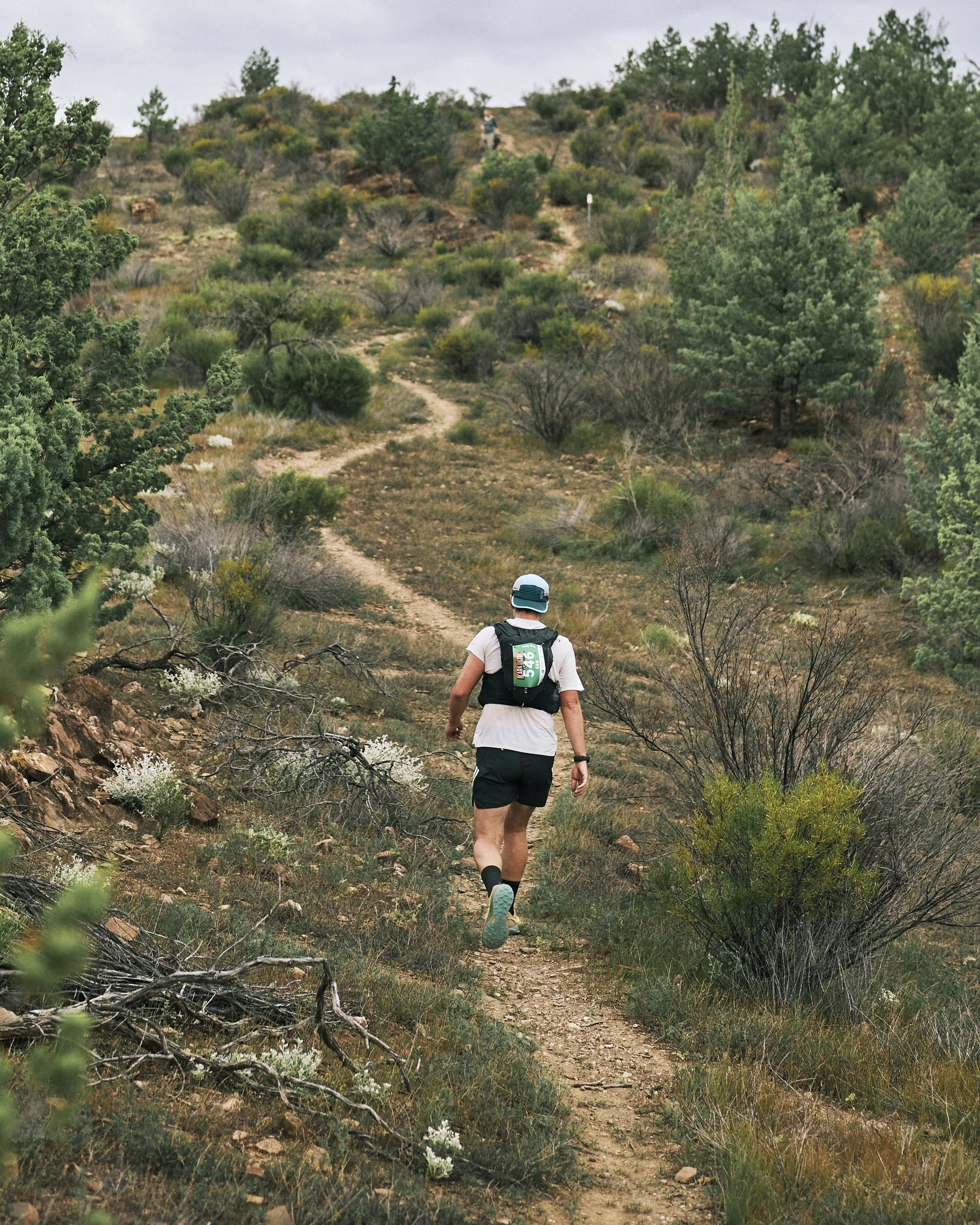 Person wearing a white t-shirt, black shorts, and a hat, hiking on a dirt trail surrounded by green bushes and trees in a hilly terrain.