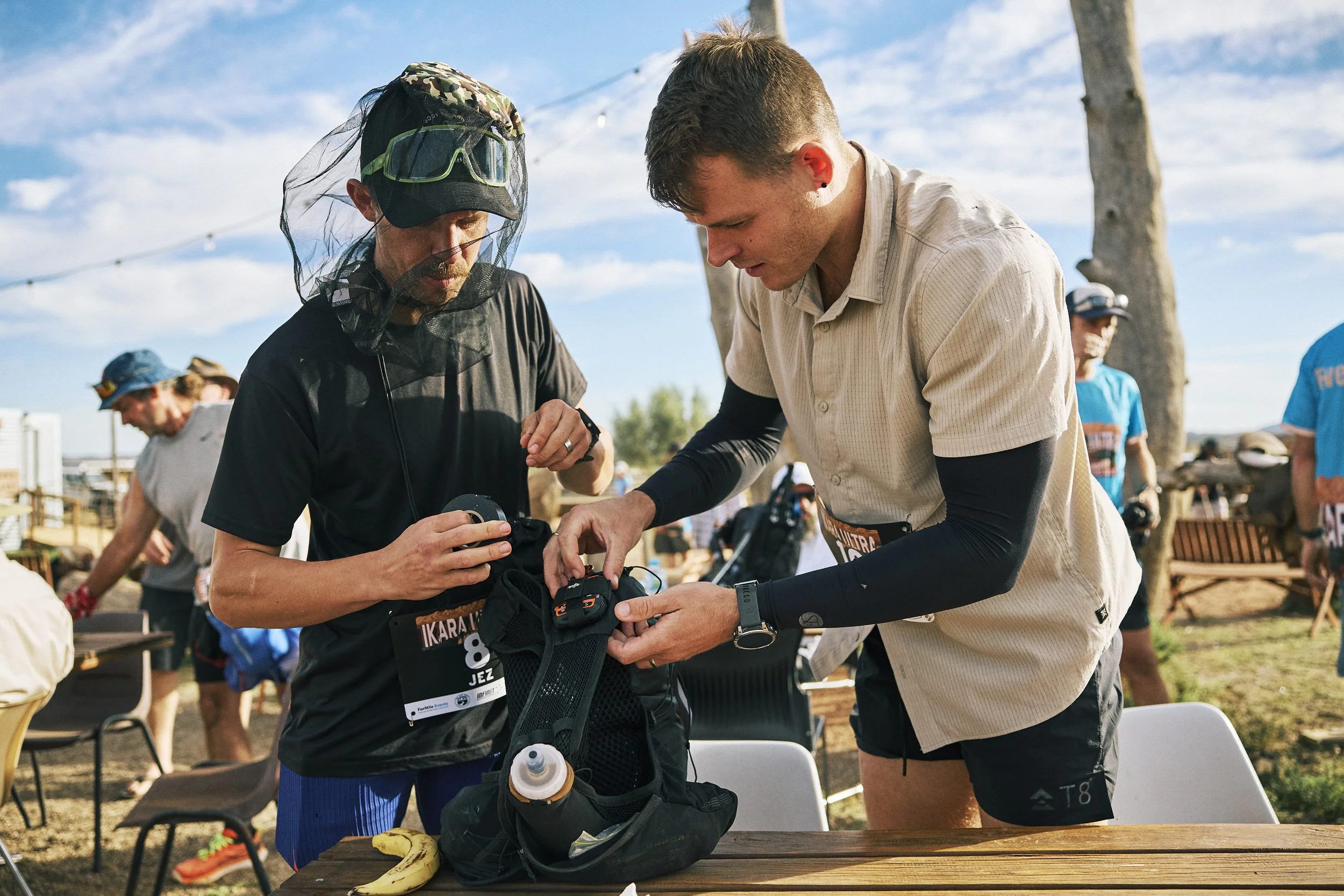 Two men are preparing for a race outdoors with trees and other people in the background. One man is wearing a black shirt, a black hat with a green visor, and a veil, while the other is in a beige shirt and black shorts. They are organizing their gea