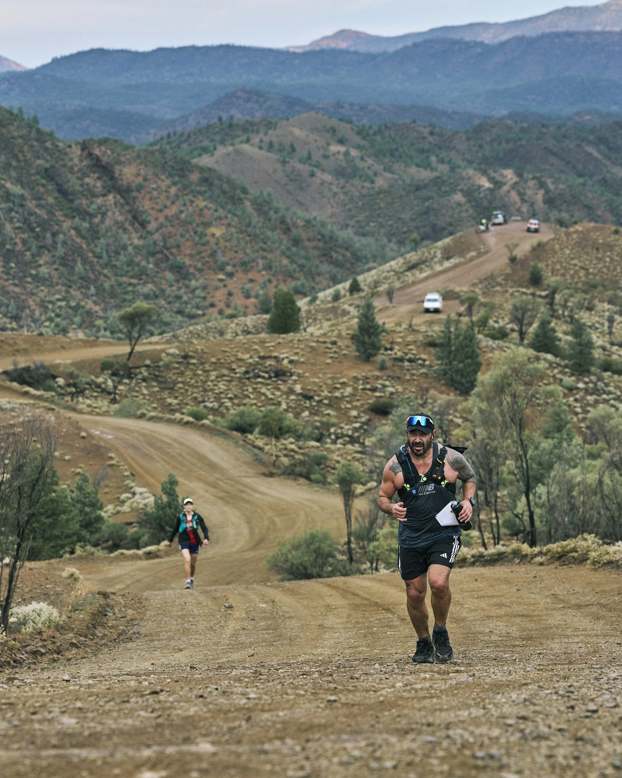 Two people trail running on a dirt path in a mountainous landscape, with one man in the foreground wearing a black shirt and shorts, and a woman further behind, with mountains in the background.