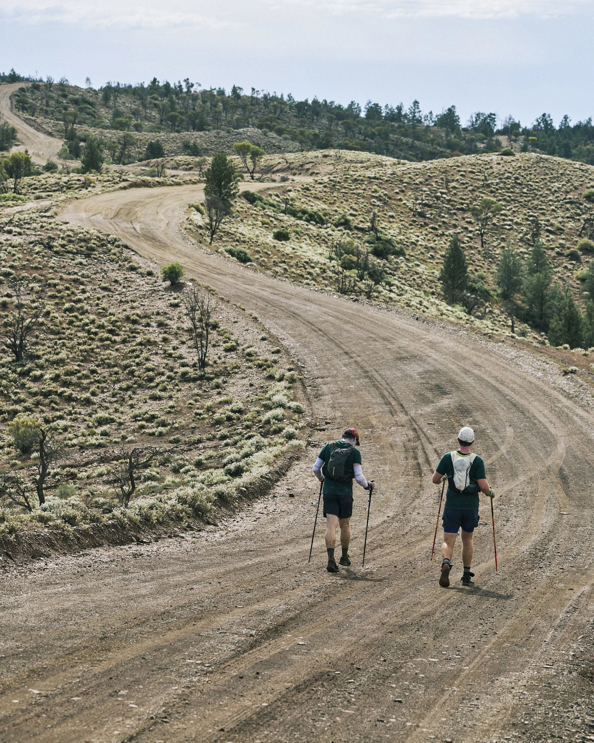 Two hikers with backpacks and hiking poles walking on a dirt trail through a semi-arid landscape with sparse vegetation and rolling hills.