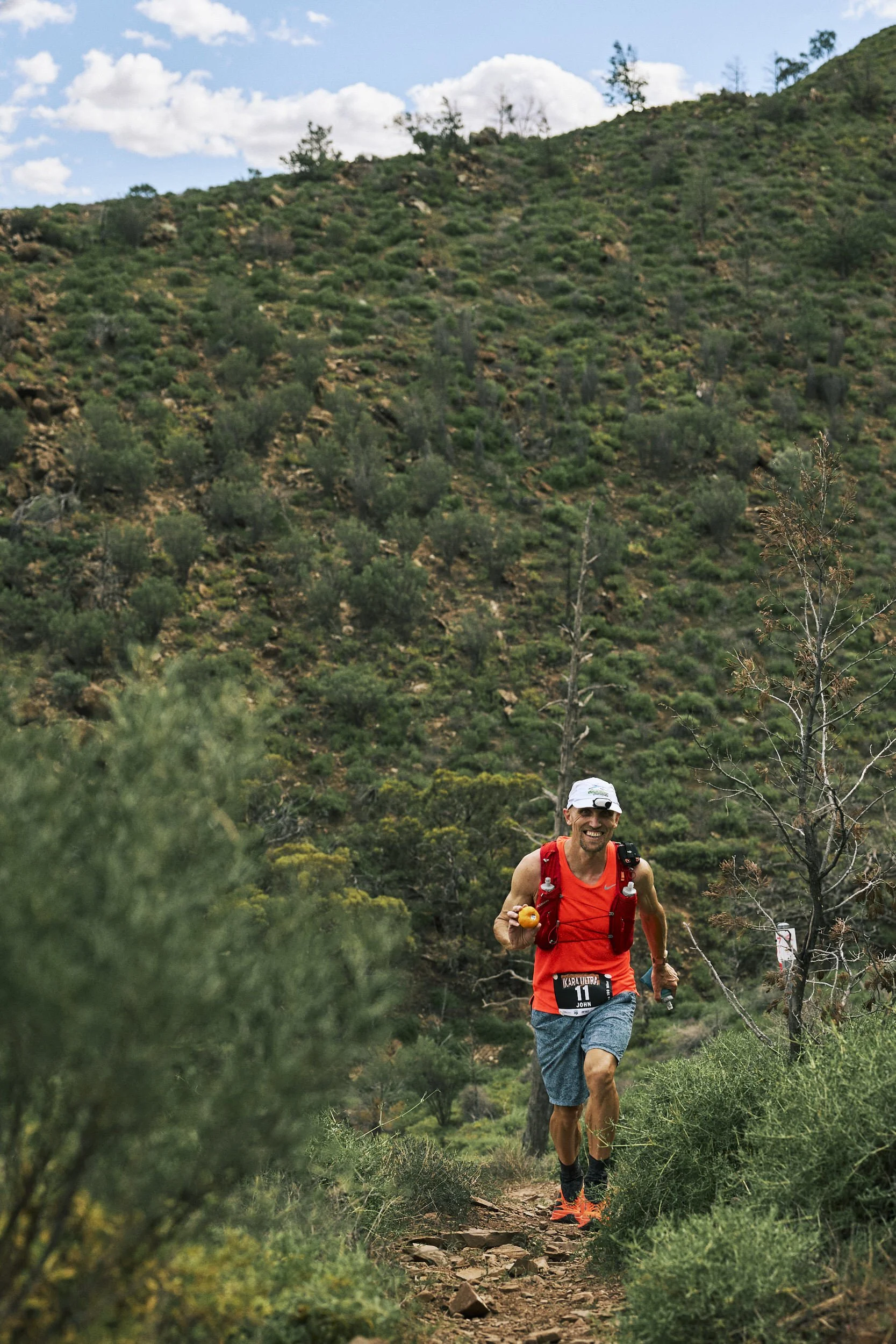 A male runner wearing a red tank top, blue shorts, and orange running shoes, trail running on a dirt path in a green mountainous area with bushes and trees.
