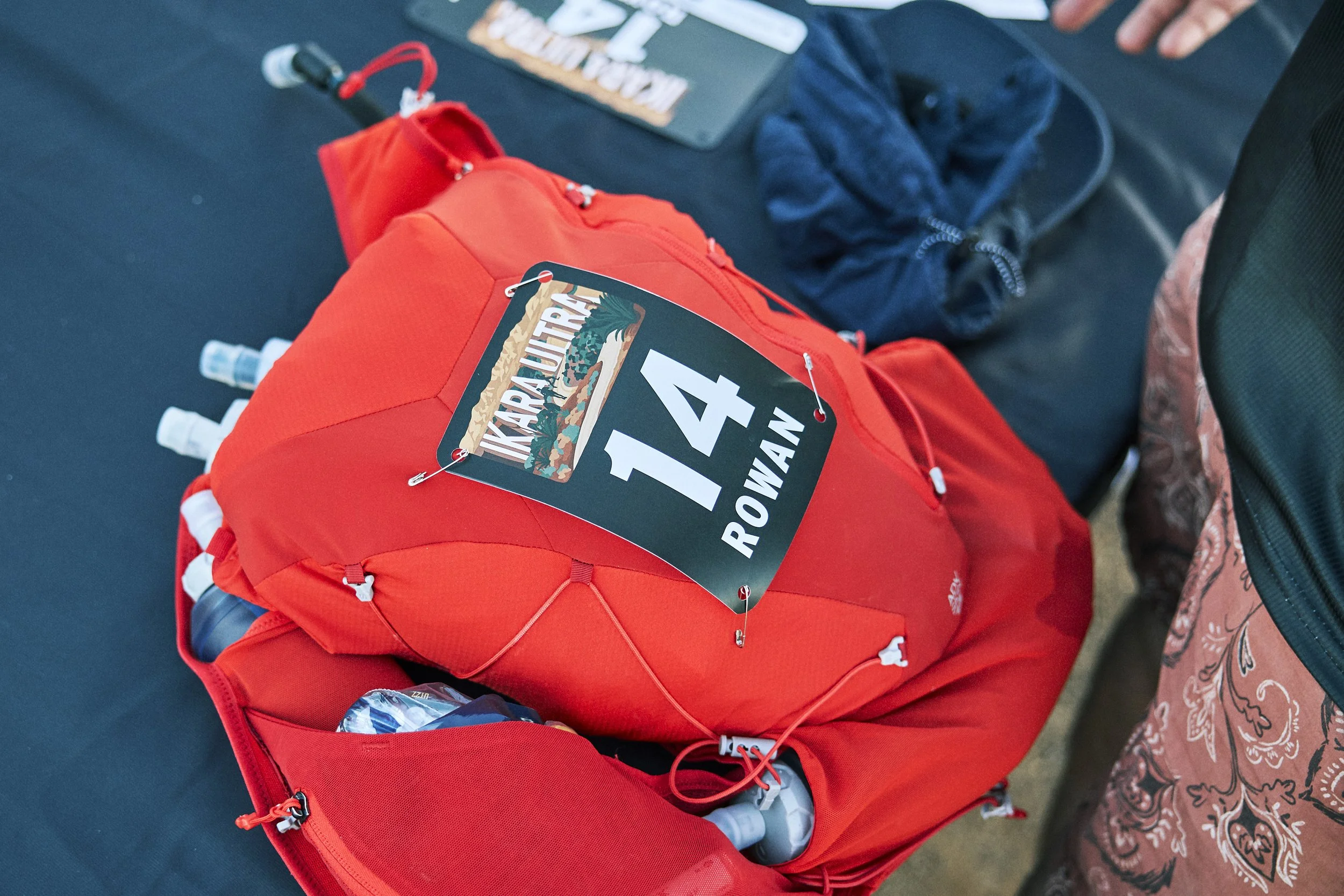 Close-up of a red backpack on a table, with a race bib labeled '14' attached to it. A hand is visible nearby, and some clothing and objects are partially visible around the backpack.