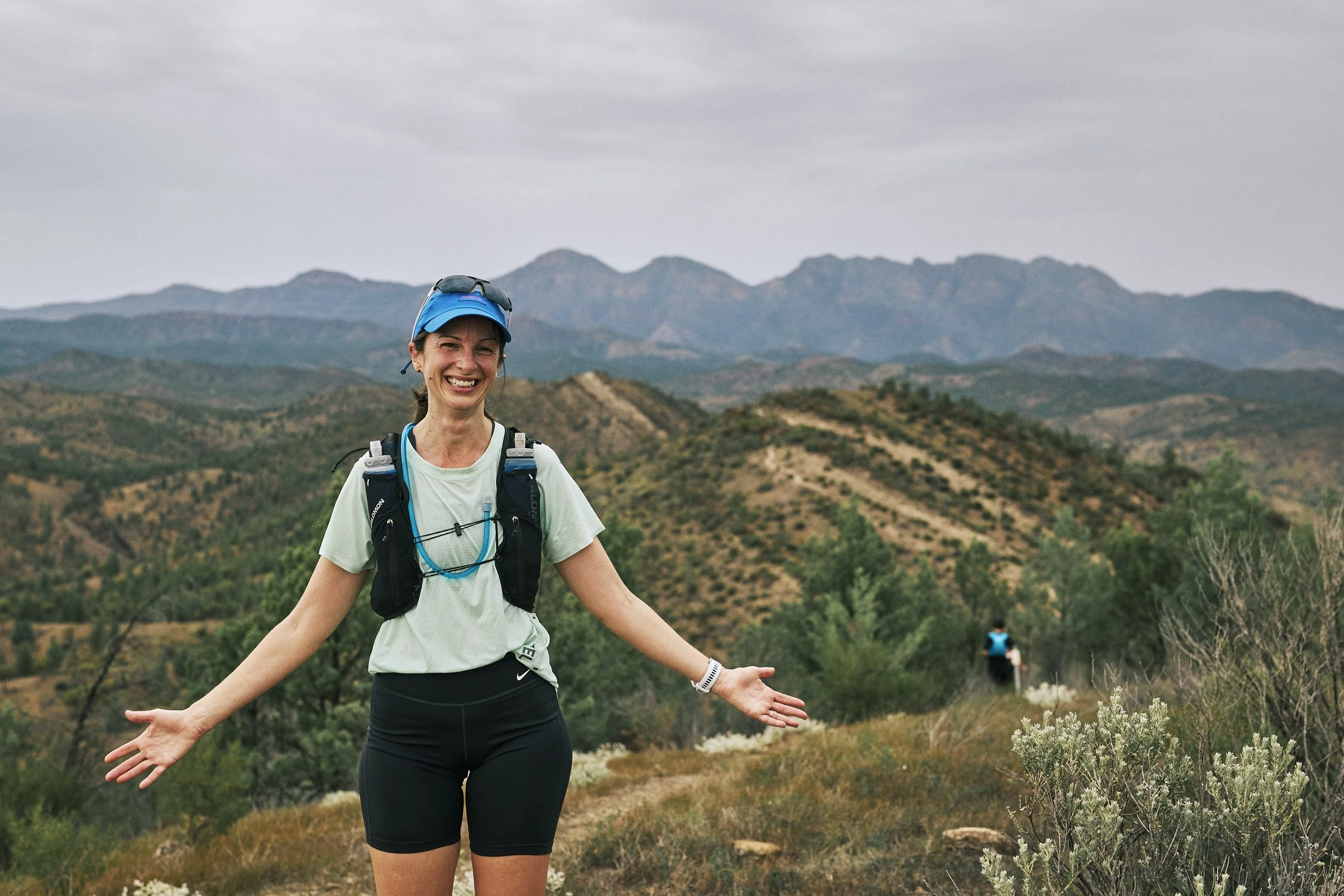 A woman with a big smile, wearing a blue cap, sunglasses on top, a white t-shirt, and black shorts, standing outdoors with her arms open wide against a background of rolling hills and mountains on a cloudy day.