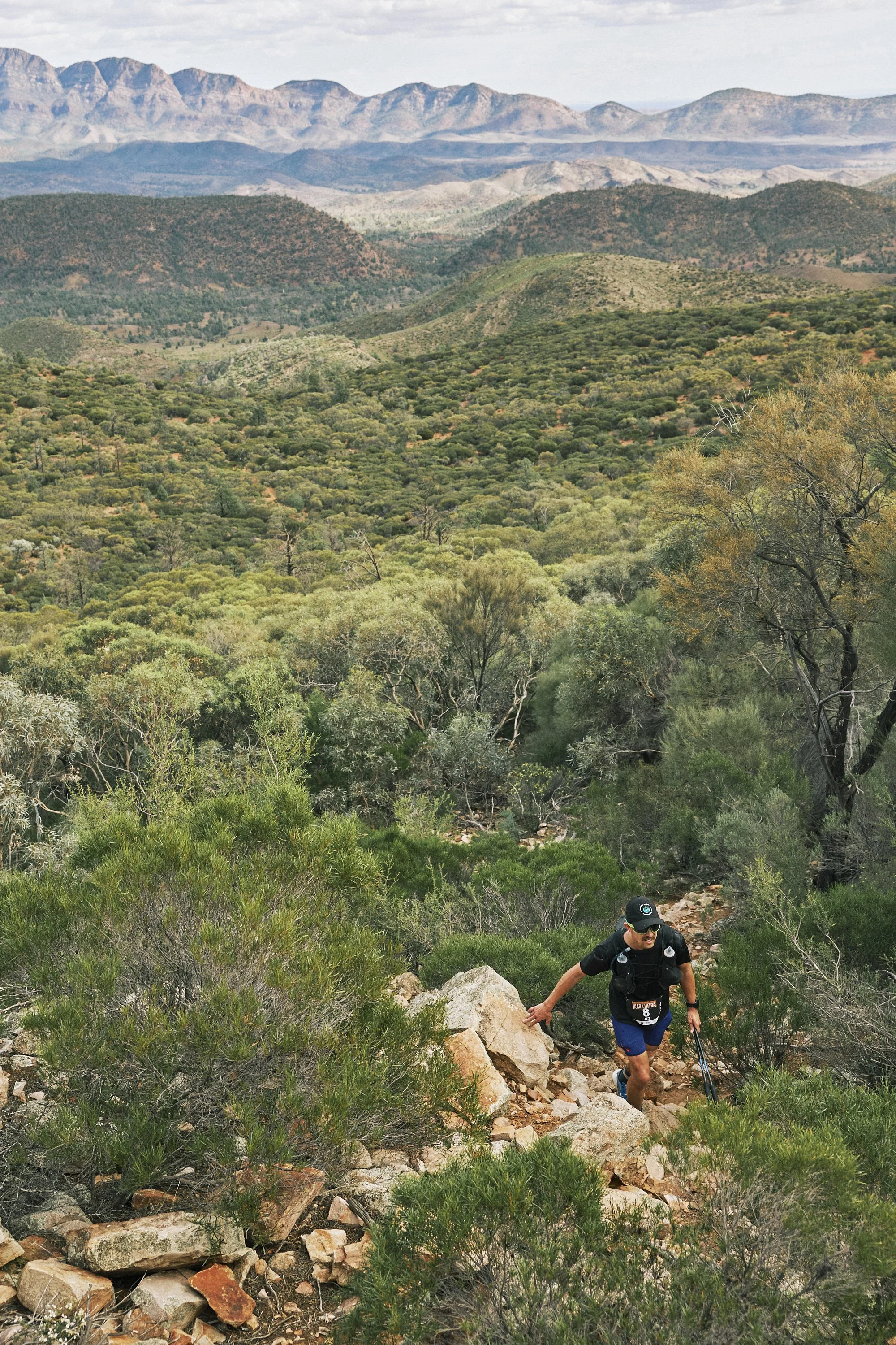 A man wearing a black shirt, blue shorts, and a cap hiking up a rocky trail in a green, mountainous landscape with distant mountains and a cloudy sky.