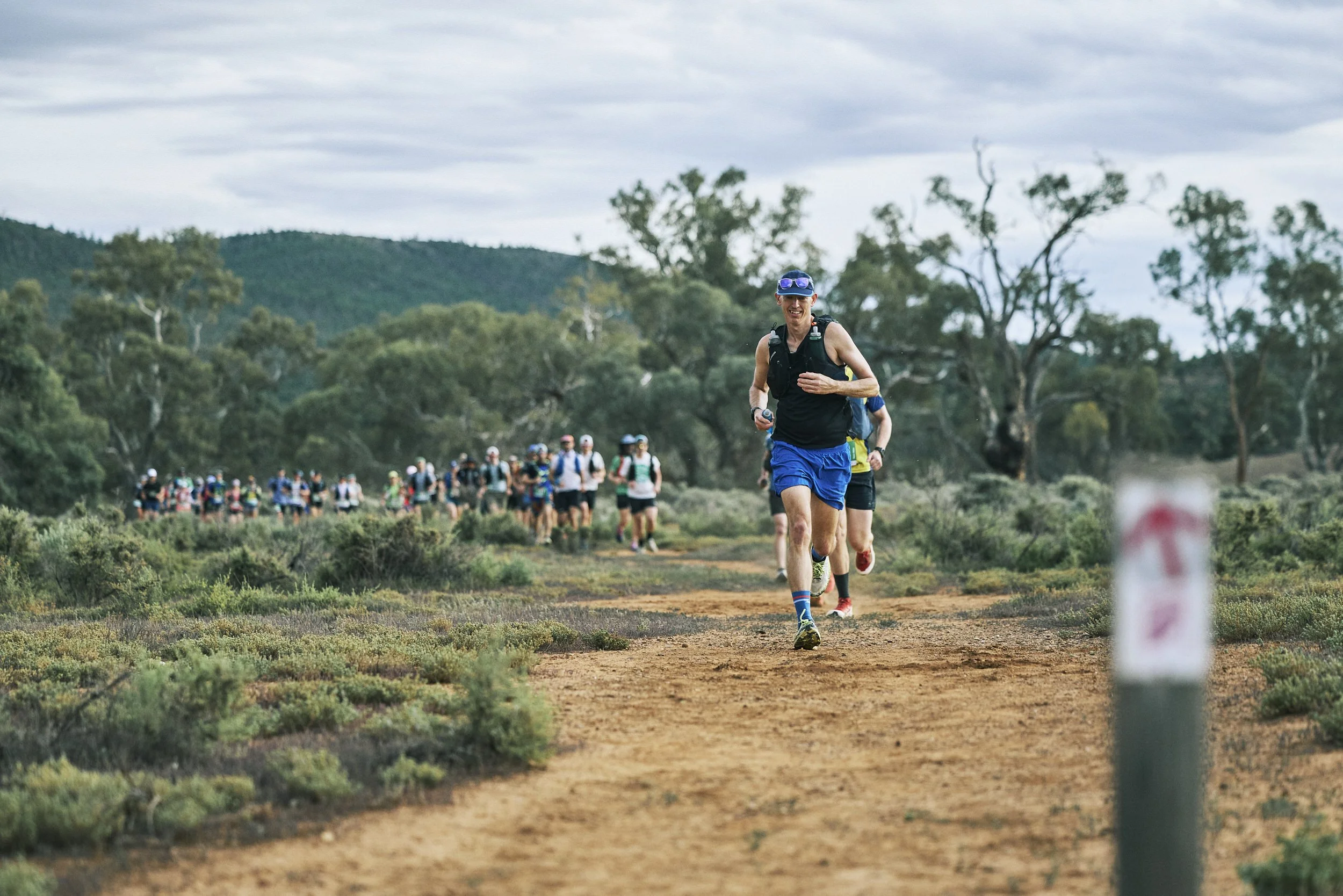 A group of people participating in a trail running event in a natural outdoor setting with green trees and hills in the background. The focus is on a smiling male runner wearing a blue cap, black sleeveless shirt, and blue shorts, leading the pack.