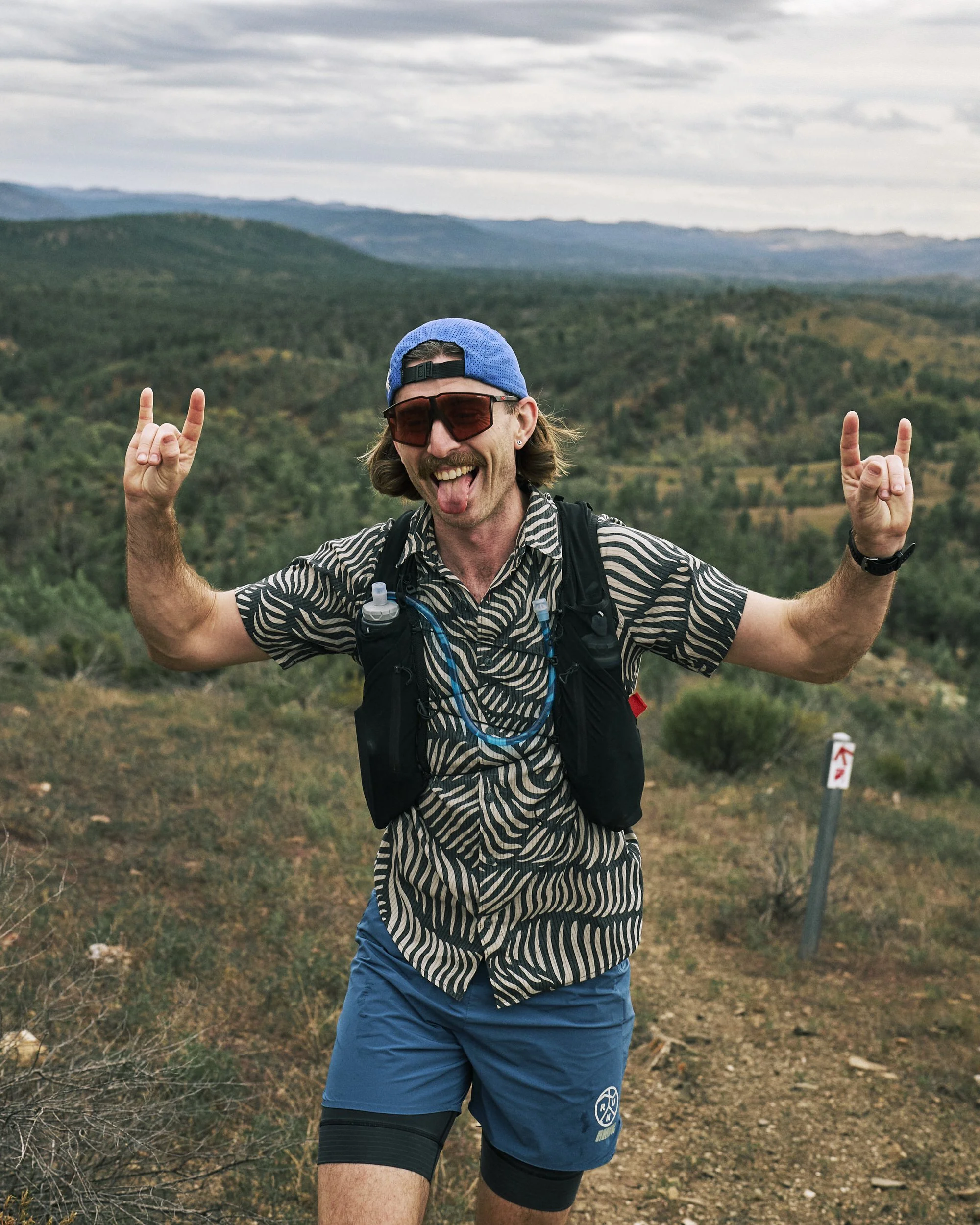 A man with long hair, wearing sunglasses, a blue cap, a zebra-striped shirt, blue running shorts, and a backpack, is making rock and roll hand gestures and sticking out his tongue while outdoors in a hilly landscape with cloudy skies.