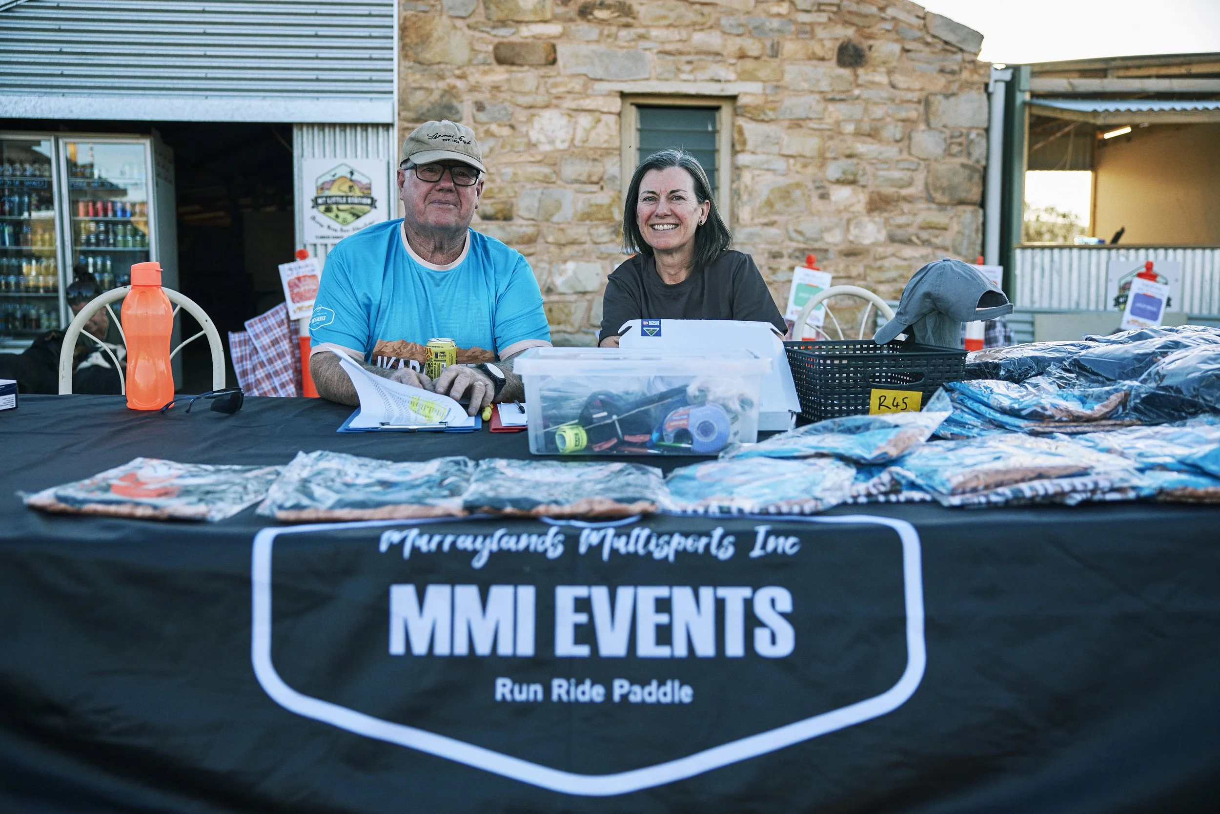 Two smiling individuals sitting at a table with a black tablecloth displaying the logo of Murraylands Multisports Inc., an event organization. The table is covered with clothes, papers, and an orange water bottle. Behind them, there's a brick buildin